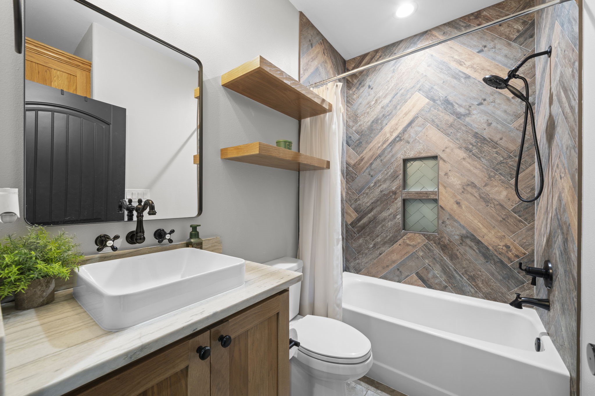 1655 Old Cuero Road Yoakum, TX 77995 - Photo 14 of 24 This stylish full bathroom blends modern farmhouse design with high-end finishes and thoughtful details. The custom wood-look herringbone tile accent wall creates a striking focal point, complemented by a built-in shower niche for convenient storage. A sleek black rainfall shower fixture and coordinating hardware add a contemporary touch, while the furniture-style vanity features a vessel sink, and matte black fixtures.