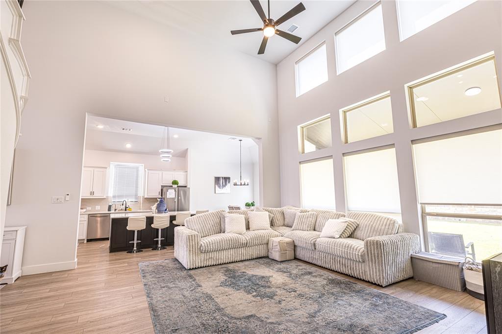 12135 Cotton Field Road Frisco, TX 75035 - Photo 5 of 38 Double-height ceiling living space featuring a contemporary ceiling fan, expansive multiple-pane windows, wood-finish flooring, and an open concept layout connecting to a kitchen with an island and pendant lighting