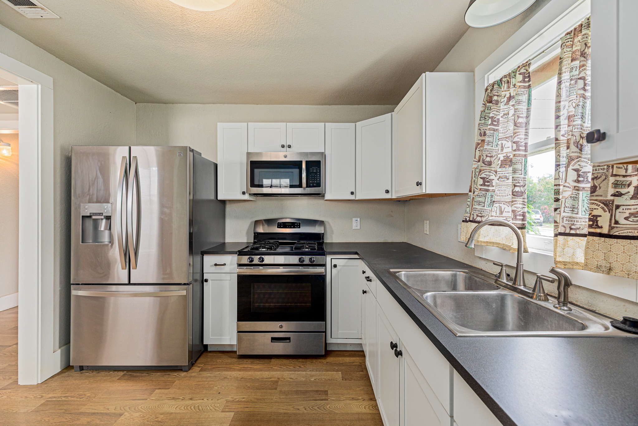 322 Blanco Avenue Luling, TX 78648 - Photo 9 of 20 A compact kitchen with white cabinetry and dark countertops, featuring essential appliances and a double-basin sink beneath a window.
