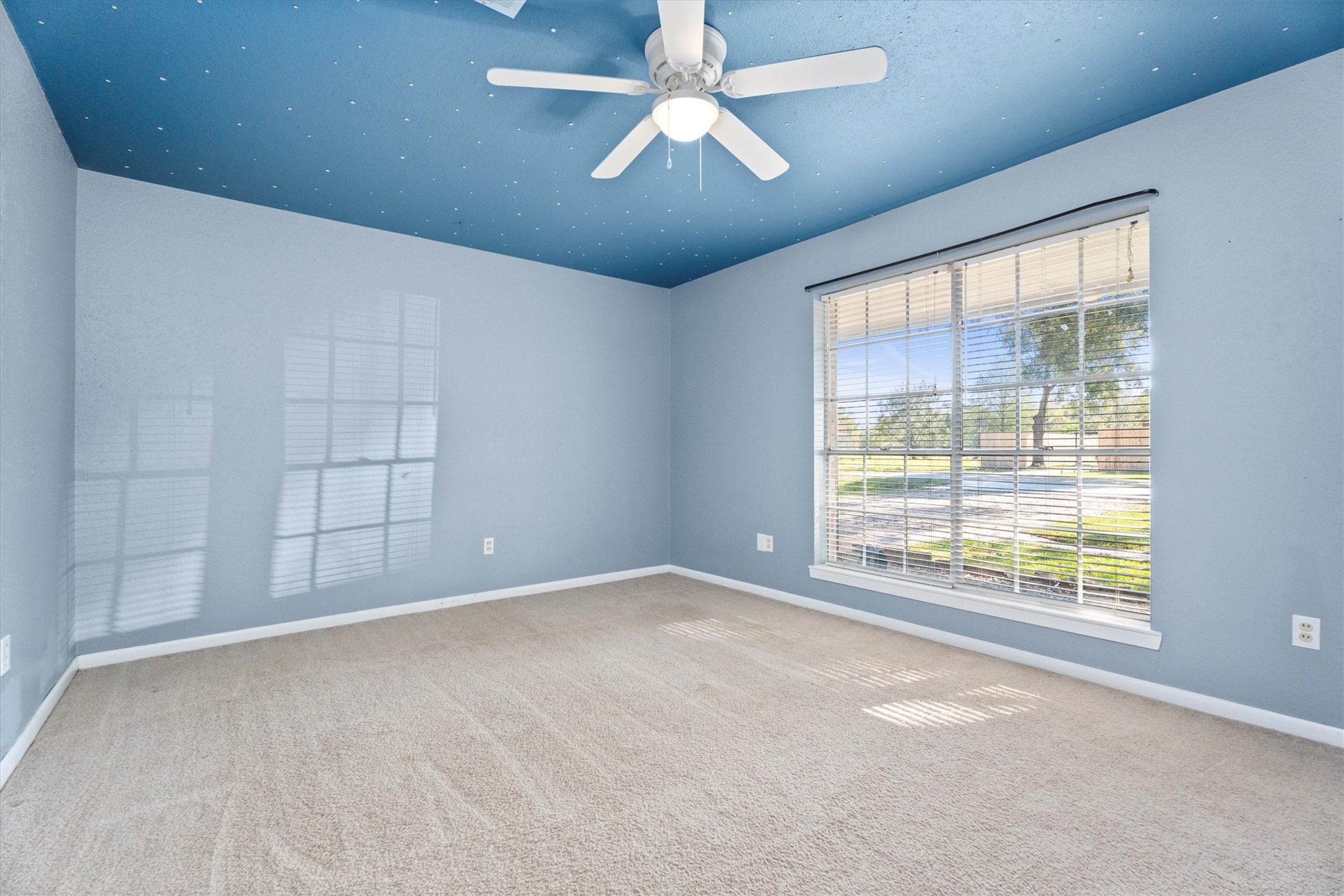 104 Anna Street Tomball, TX 77375 - Photo 15 of 32 The third bedroom off to the right of the entry with a large window at the front of the house has same neutral carpet, and a unique darker blue ceiling featuring small white stars. Equipped with a ceiling fan for comfort. There's a closet to the right that is not shown in the picture.