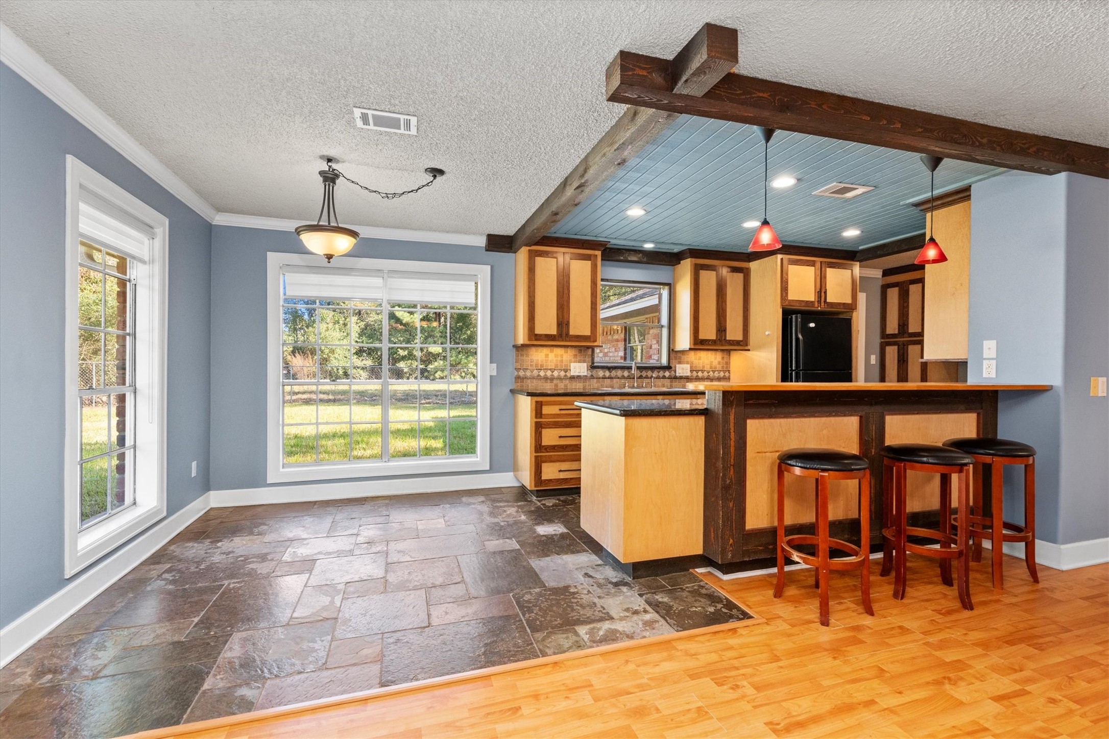 104 Anna Street Tomball, TX 77375 - Photo 5 of 32 Bright and inviting open kitchen and dining area with large windows, modern pendant lighting, and a cozy breakfast bar. Custom cabinetry features a mix of wood tones and under-cabinet lighting. Slate tile flooring and rustic-looking, exposed, faux beams create a warm, stylish ambiance.
