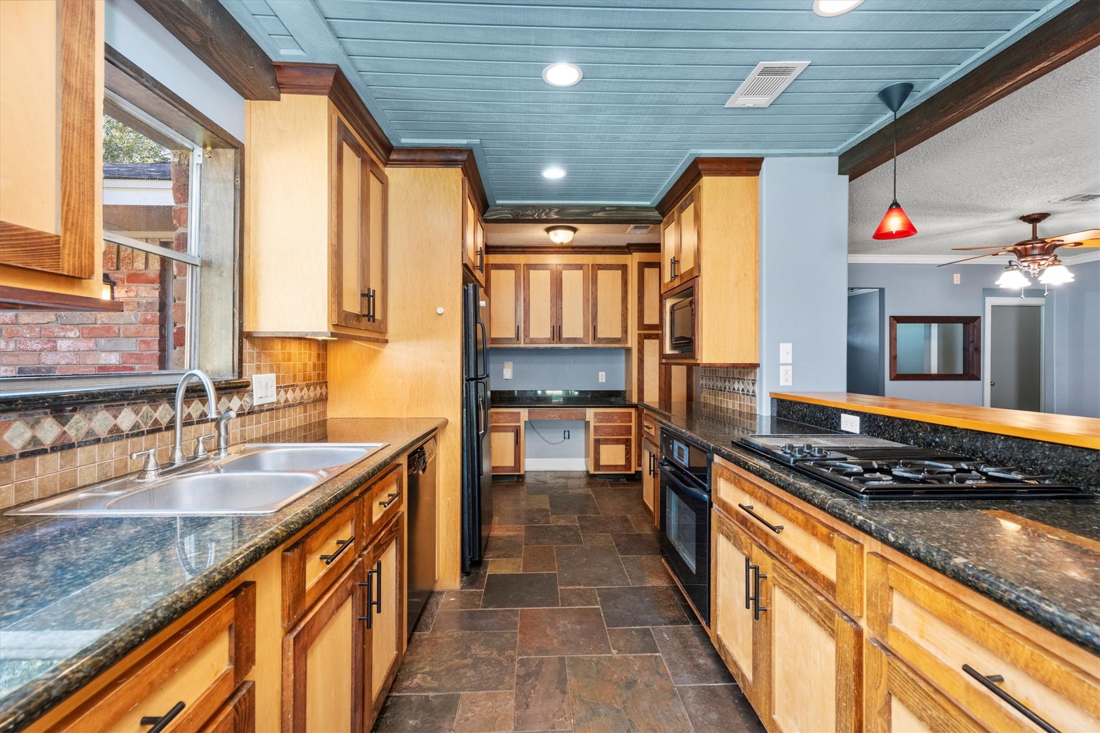 104 Anna Street Tomball, TX 77375 - Photo 6 of 32 This is a spacious kitchen featuring granite countertops, custom wooden cabinetry, and a tiled backsplash. It includes modern appliances, a double sink, and a gas cooktop with grill. The room has a mix of earthy tones with a touch of blue on the ceiling and is well-lit with both natural (window over sink) and recessed lighting.