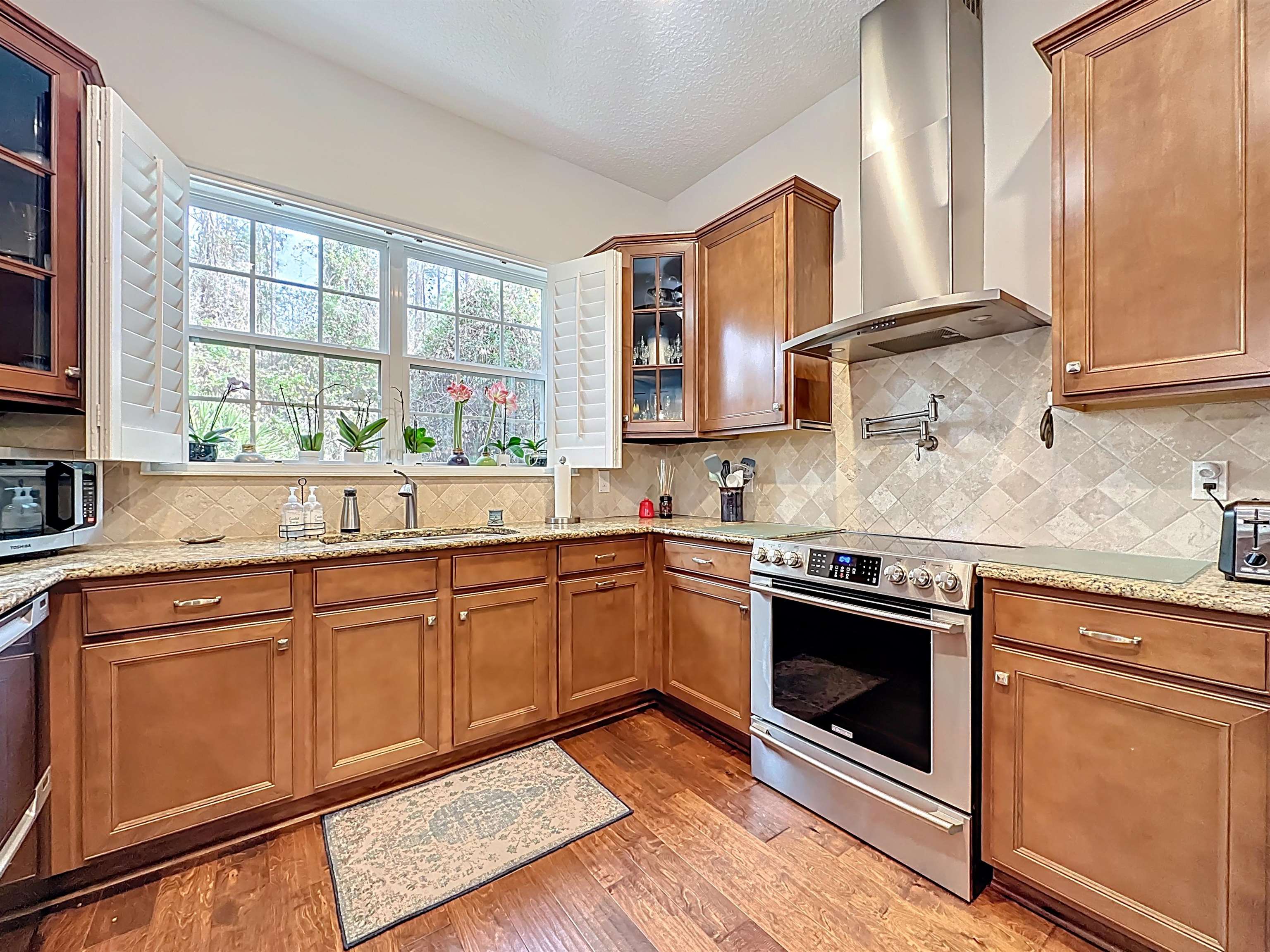 130 Grand Ravine Drive St. Augustine, FL 32086 - Photo 14 of 42 Kitchen with brown cabinets, glass insert cabinets, wall chimney range hood, appliances with stainless steel finishes, and light stone countertops