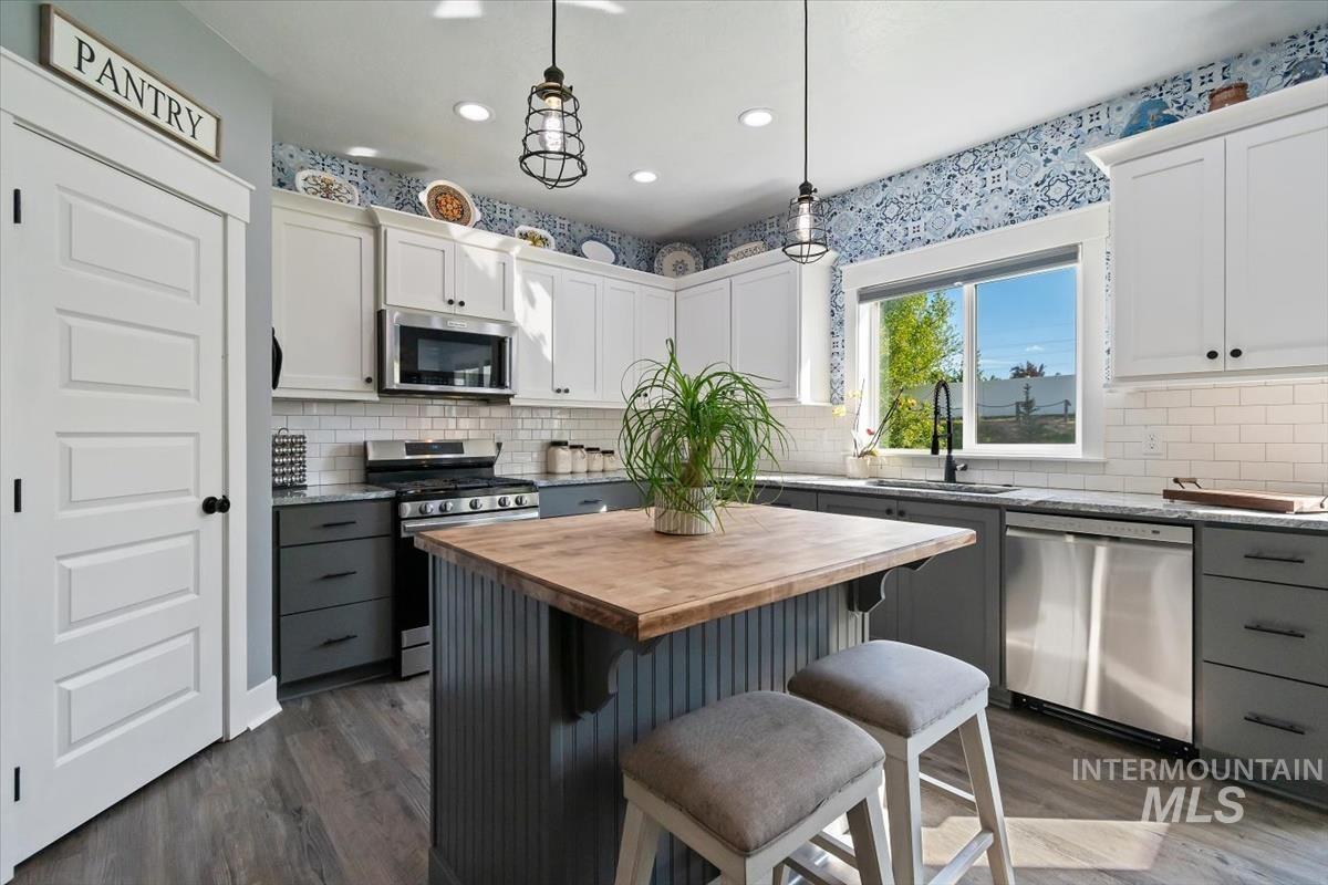 15015 Leon Lane Caldwell, ID 83607 - Photo 5 of 44 Contemporary kitchen featuring wood-finish flooring, two-tone cabinetry with white upper and dark gray lower cabinets, a subway tile backsplash, stainless steel appliances, and a central island with a wood countertop