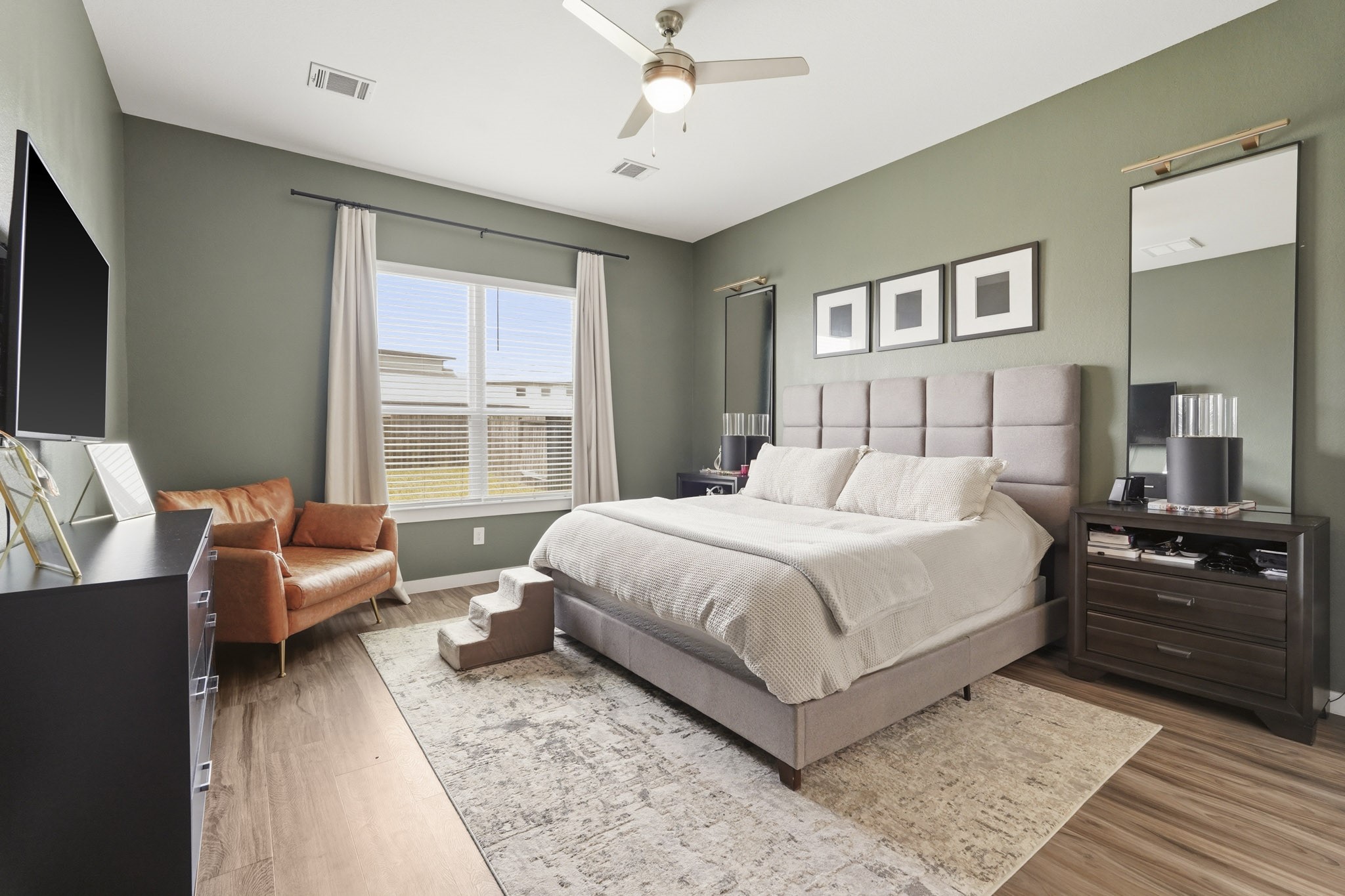 22522 Modica Rift Lane Katy, TX 77449 - Photo 20 of 38 Your personal sanctuary awaits. Wrapped in a calming sage green that feels both grounding and refreshing, the spacious primary bedroom offers everything you need to rest, recharge, and feel at home. A tall upholstered panel headboard anchors the room, flanked by nightstands, layered mirrors, and warm accent lighting. A cognac leather accent chair tucks into the corner — the perfect spot for morning coffee or a quiet evening read. This is a bedroom designed to feel like a retreat every single night.