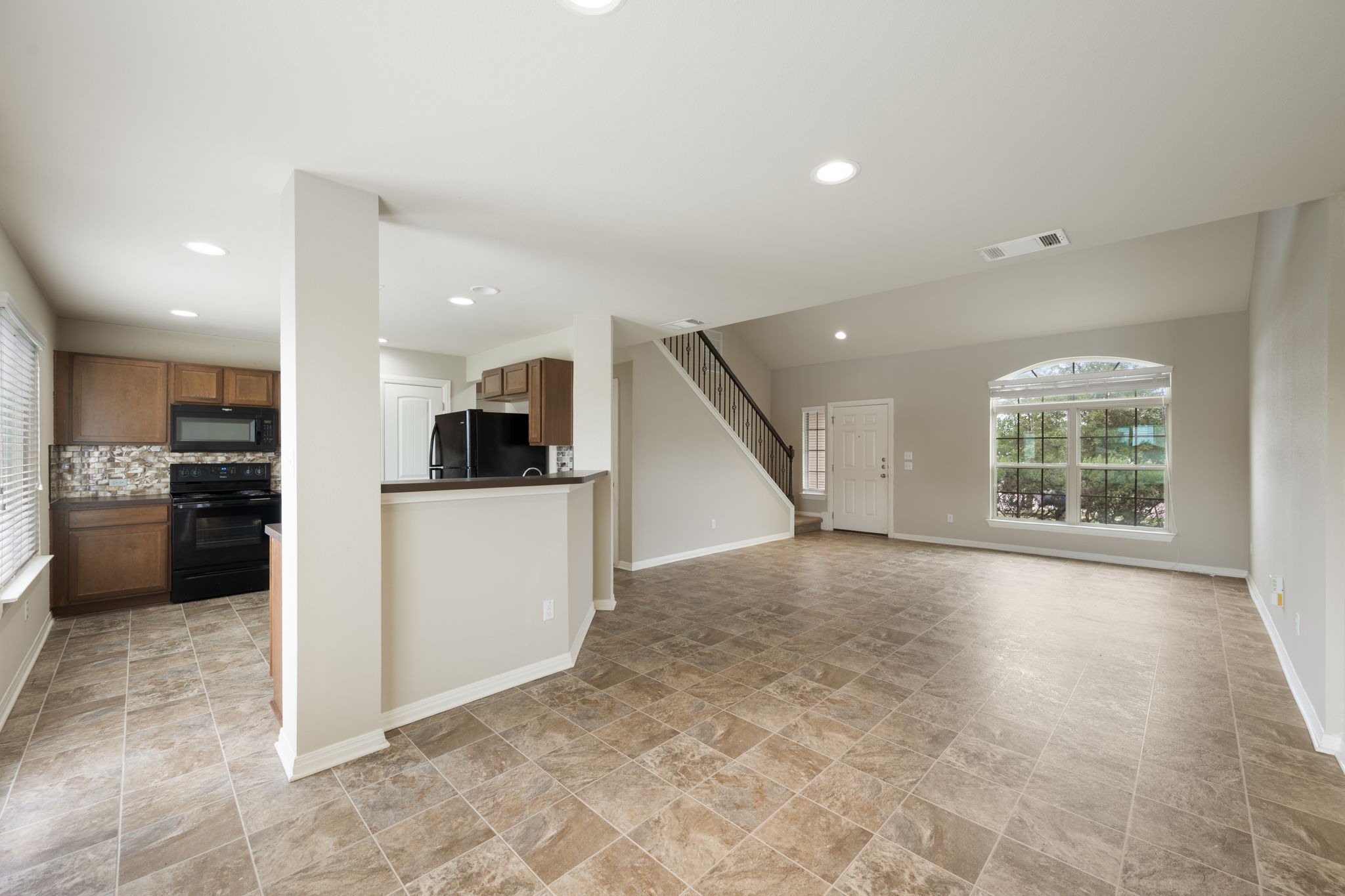 12701 Carillon Way Manor, TX 78653 - Photo 4 of 20 Open-concept living area featuring tile flooring, a kitchen with dark cabinetry and black appliances, a prominent arched window, and a staircase with dark railing
