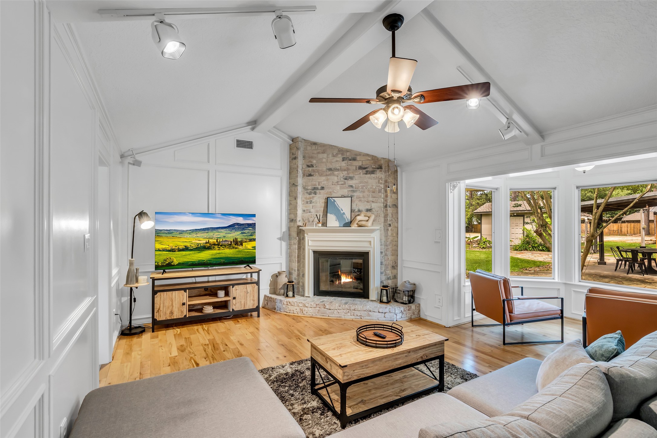 22502 Fields Lane Spring, TX 77389 - Photo 4 of 39 This inviting living room features a vaulted ceiling with a fan, a cozy brick fireplace, and abundant natural light from large windows. The space is complemented by hardwood floors, offering a blend of warmth and style with a view of the backyard.