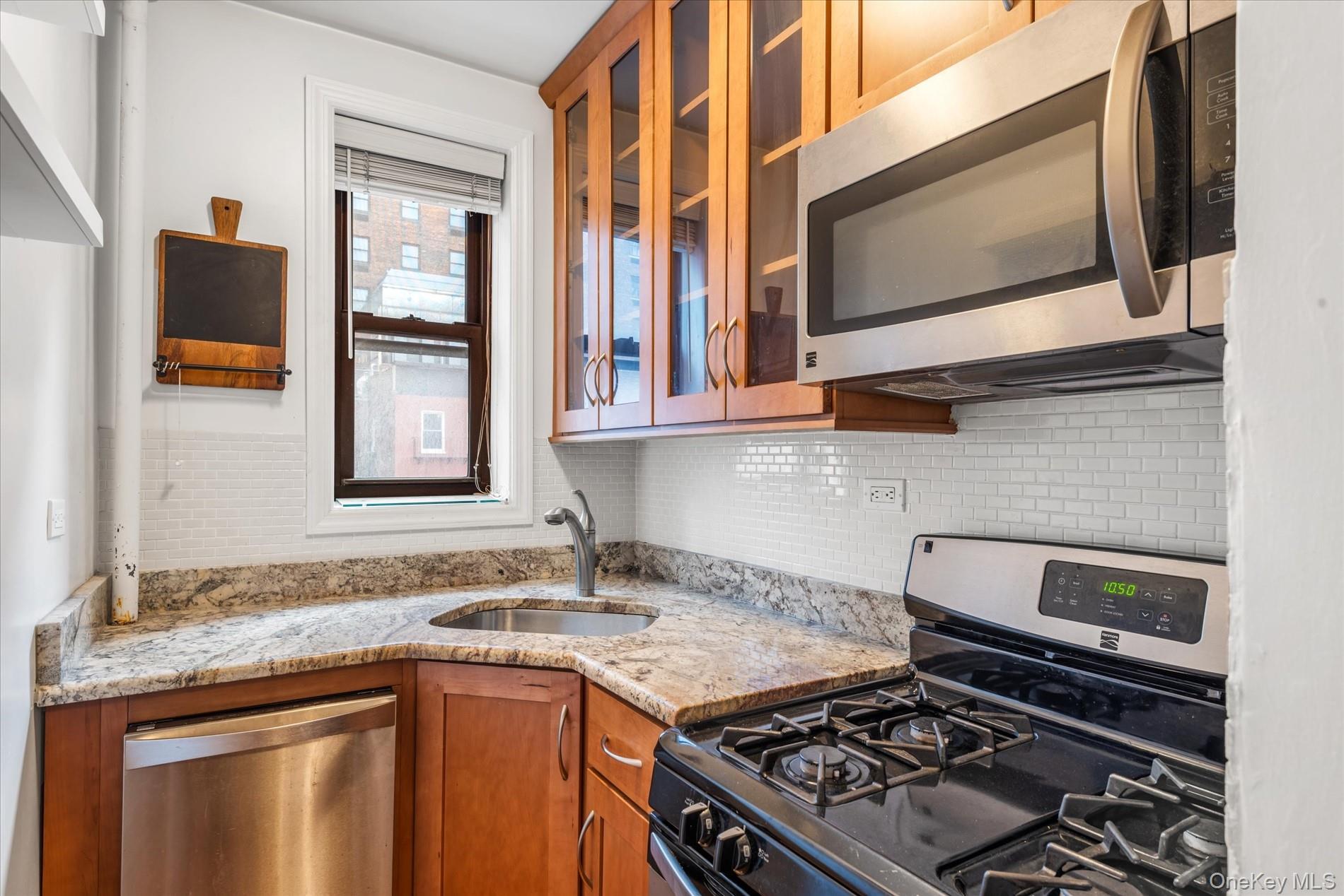 58-th Street West 400th, Unit 5G Manhattan, NY 10019 - Photo 7 of 18 Kitchen featuring brown cabinets, a sink, backsplash, appliances with stainless steel finishes, and glass insert cabinets