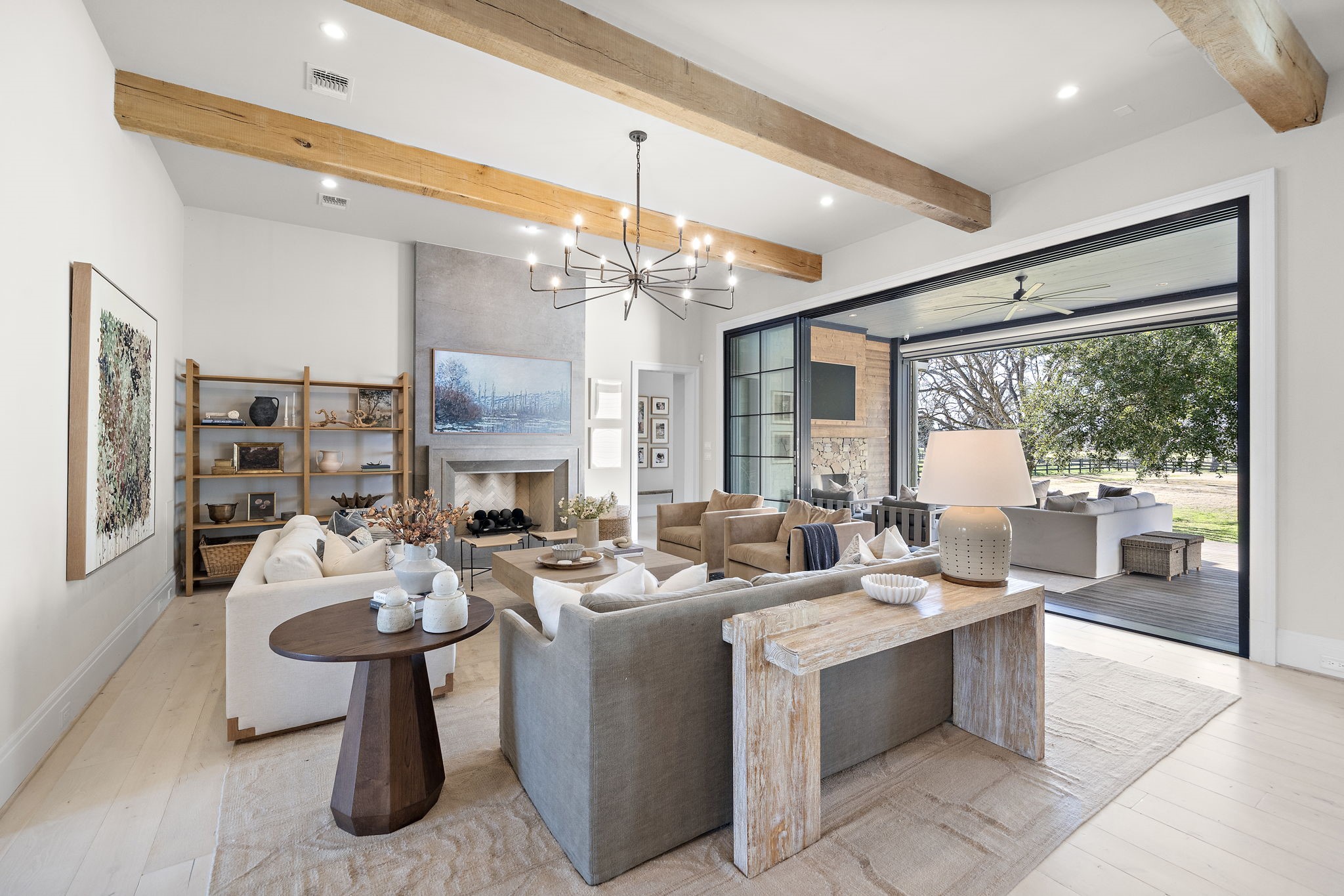 5511 Fulbrook Road Fulshear, TX 77441 - Photo 15 of 50 This spacious living room features exposed wooden beams, a modern chandelier, and a cozy fireplace. It seamlessly connects to an outdoor patio through large sliding glass doors, offering a picturesque view.