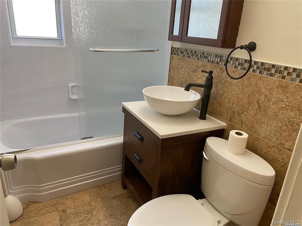 107 Baker Hill Road Great Neck, NY 11023 - Photo 12 of 33 First floor Bathroom with combined bath / shower with glass door, tile walls, vanity, and a wainscoted wall