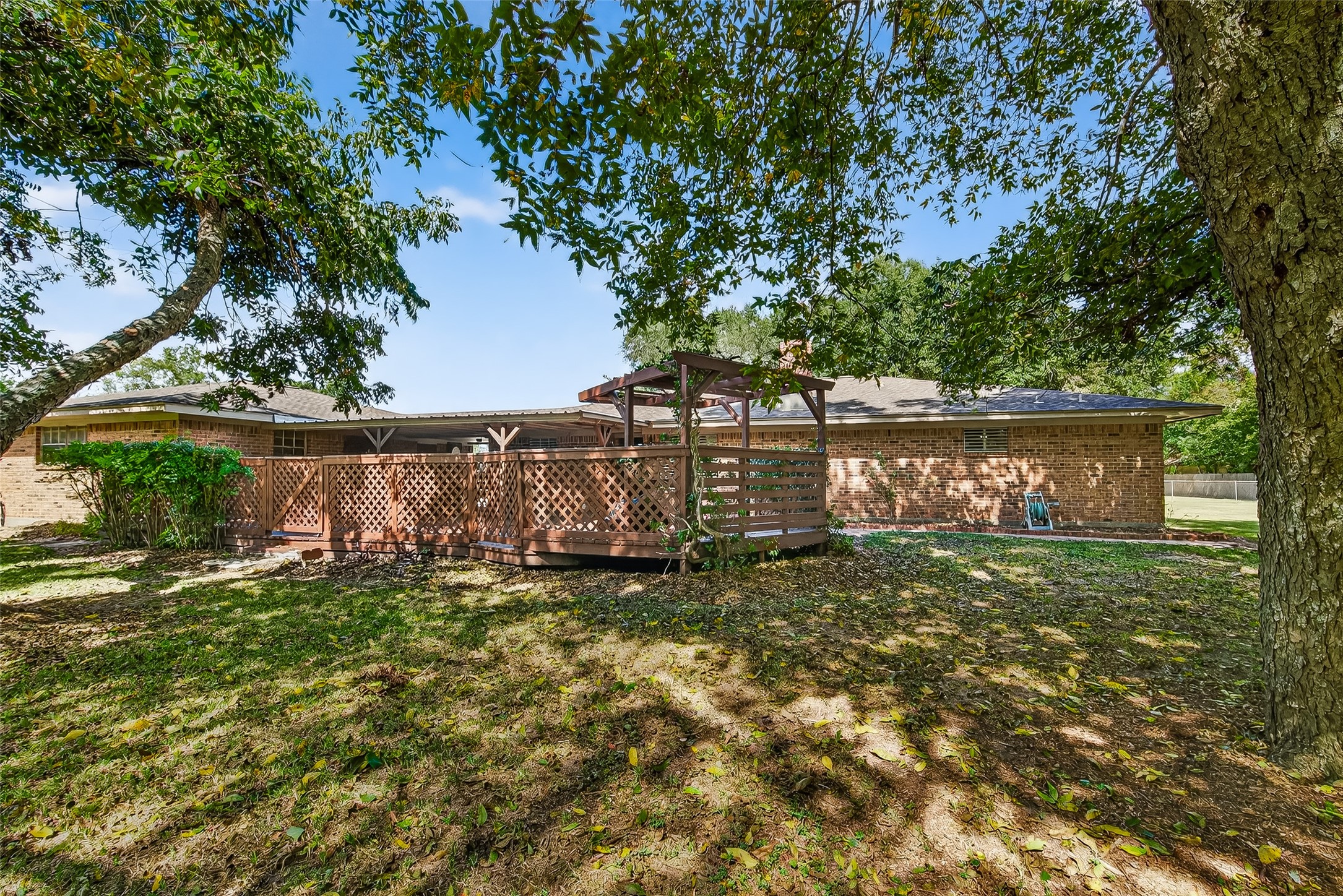 12021 7th 1/2 Street Santa Fe, TX 77510 - Photo 9 of 49 This photo showcases a backyard with a spacious wooden deck and lattice fencing, surrounded by mature trees providing shade. The brick exterior of the house complements the natural setting, creating a cozy and inviting outdoor space.