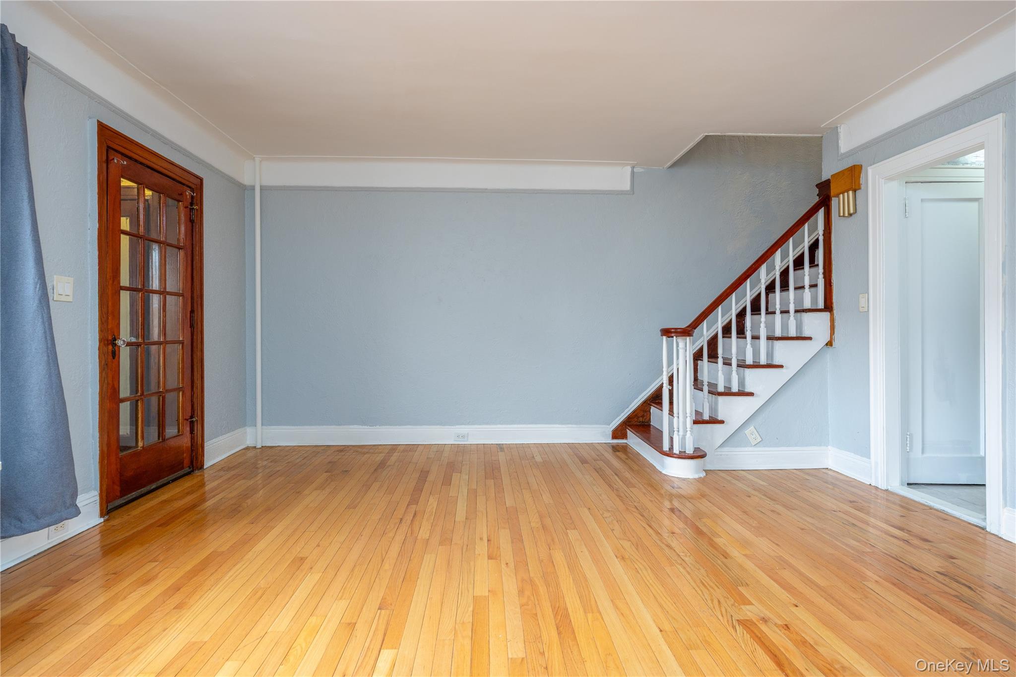 179-65 Selover Road Queens, NY 11434 - Photo 6 of 33 Living Room17'7" x 14'4"Spacious living area featuring light wood-finish hardwood flooring throughout, a corner fireplace with a stone firebox and painted brick surround, light blue wall paint, white crown molding, paneled windows, a glass-paneled entry door, and an arched doorway leading to an additional room. The space also includes a detailed staircase with white balusters, white risers, and dark wood treads