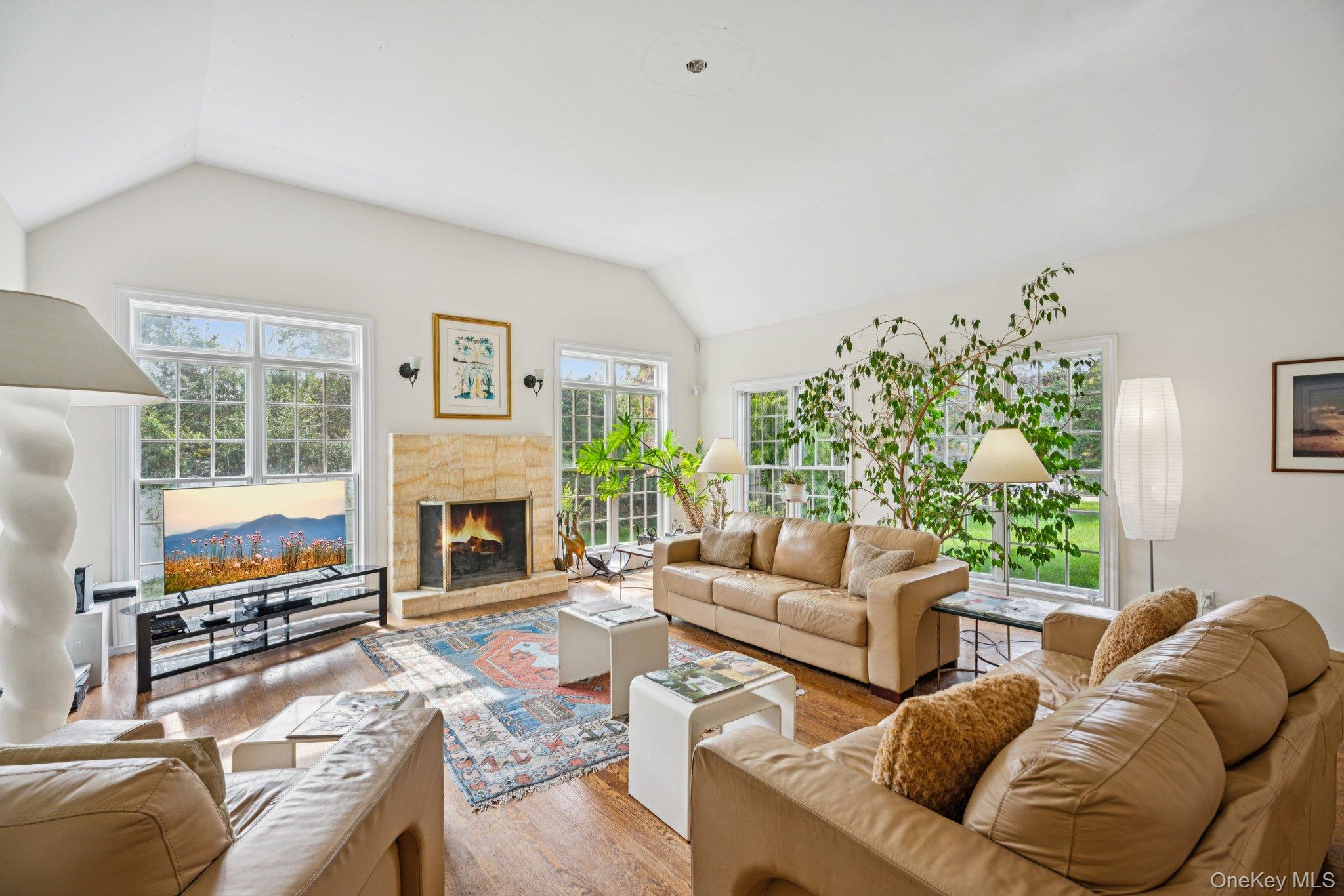 12 Laila Lane Remsenburg, NY 11960 - Photo 6 of 50 An exquisite living area showcasing vaulted ceilings, rich oak hardwood floors, and a timeless wood-burning fireplace, beautifully encased in floor-to-ceiling windows and sleek glass sliders that invite an abundance of natural light.