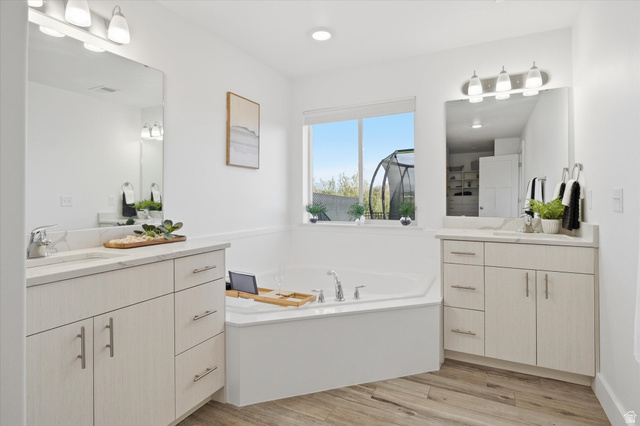 1422 West 3410th Street South Logan, UT 84321 - Photo 17 of 33 Spacious bathroom featuring a built-in soaking tub under a window, dual vanity sinks with light-toned cabinetry, coordinated countertops, and wood-finish flooring