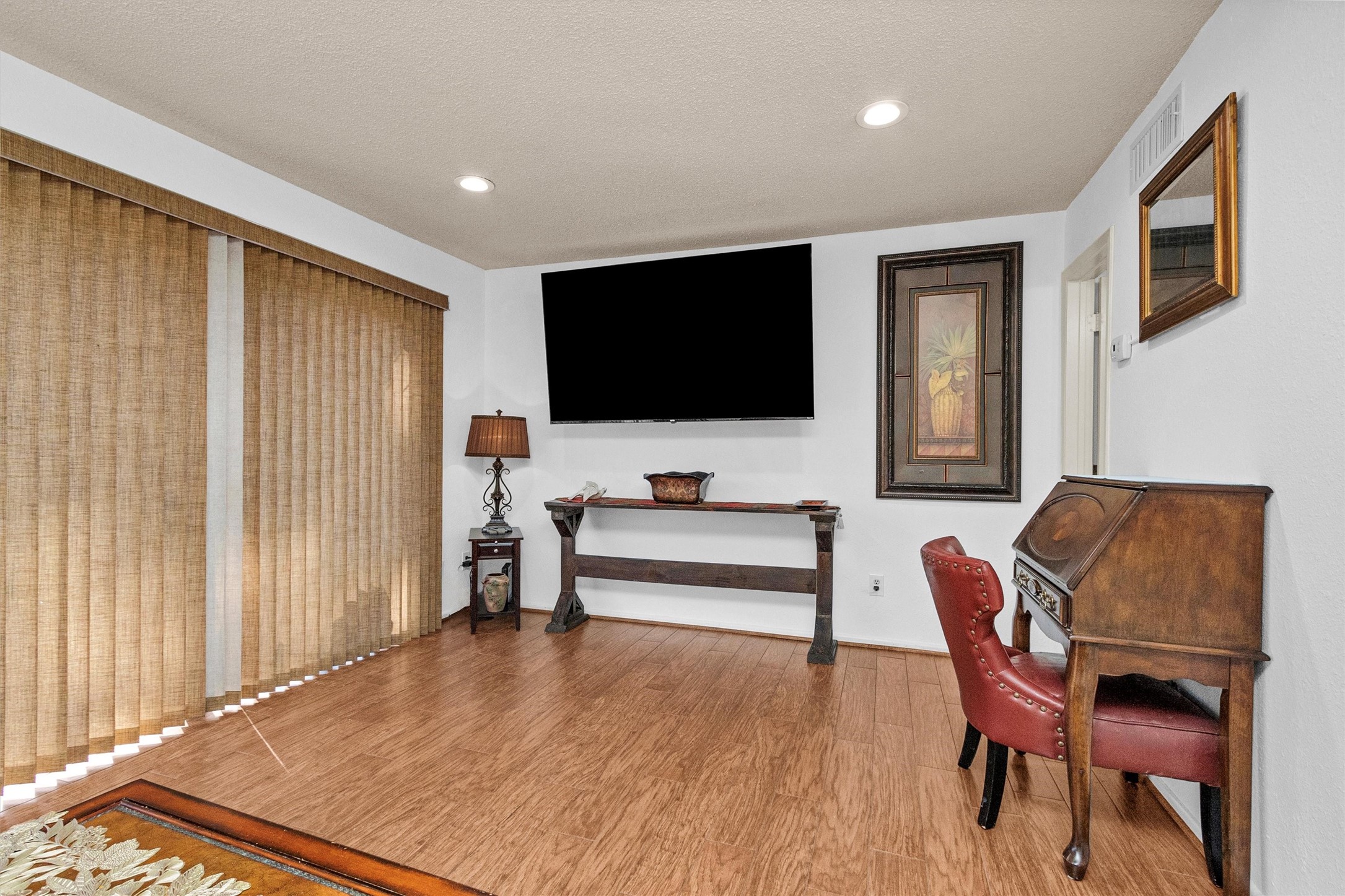12900 Walden Road, Unit D 411 Montgomery, TX 77356 - Photo 7 of 18 This inviting living space boasts warm wood flooring, a large wall-mounted TV, stylish vertical blinds, and elegant decor with a vintage desk and classic artwork, perfect for relaxation or entertainment.