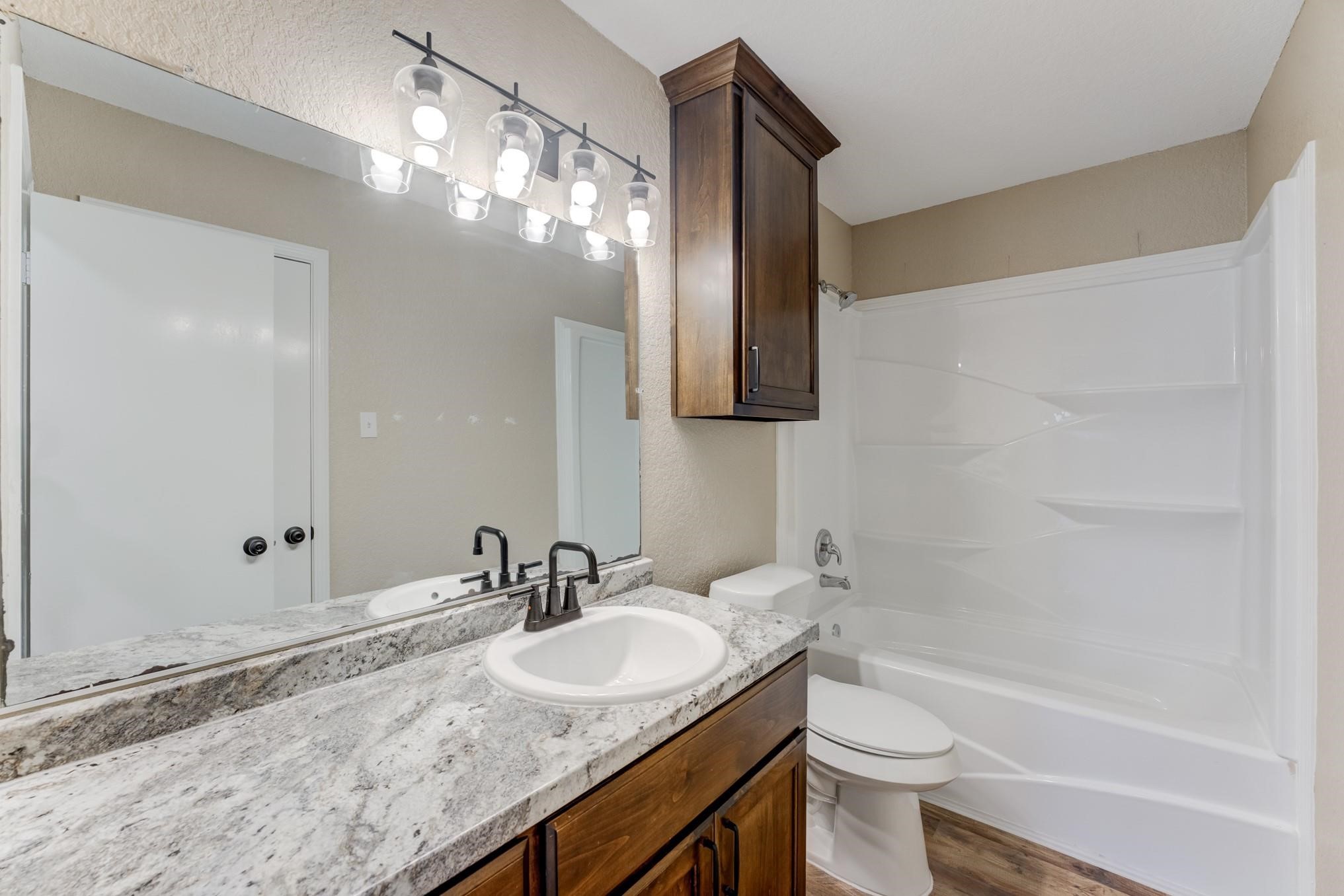 305 Yaupon Street Sweeny, TX 77480 - Photo 16 of 22 This bathroom features a modern design with a granite countertop, single sink, and dark wood cabinetry. It includes a large mirror with stylish lighting and a clean, white bathtub-shower combo. The space has a neutral color palette and functional layout.