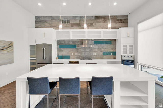 7713 Navarro View Court Midvale, UT 84047 - Photo 35 of 41 Spacious kitchen featuring a large white island with integrated shelving, stainless steel appliances, a unique tile backsplash with blue accents, white cabinetry, and pendant lighting