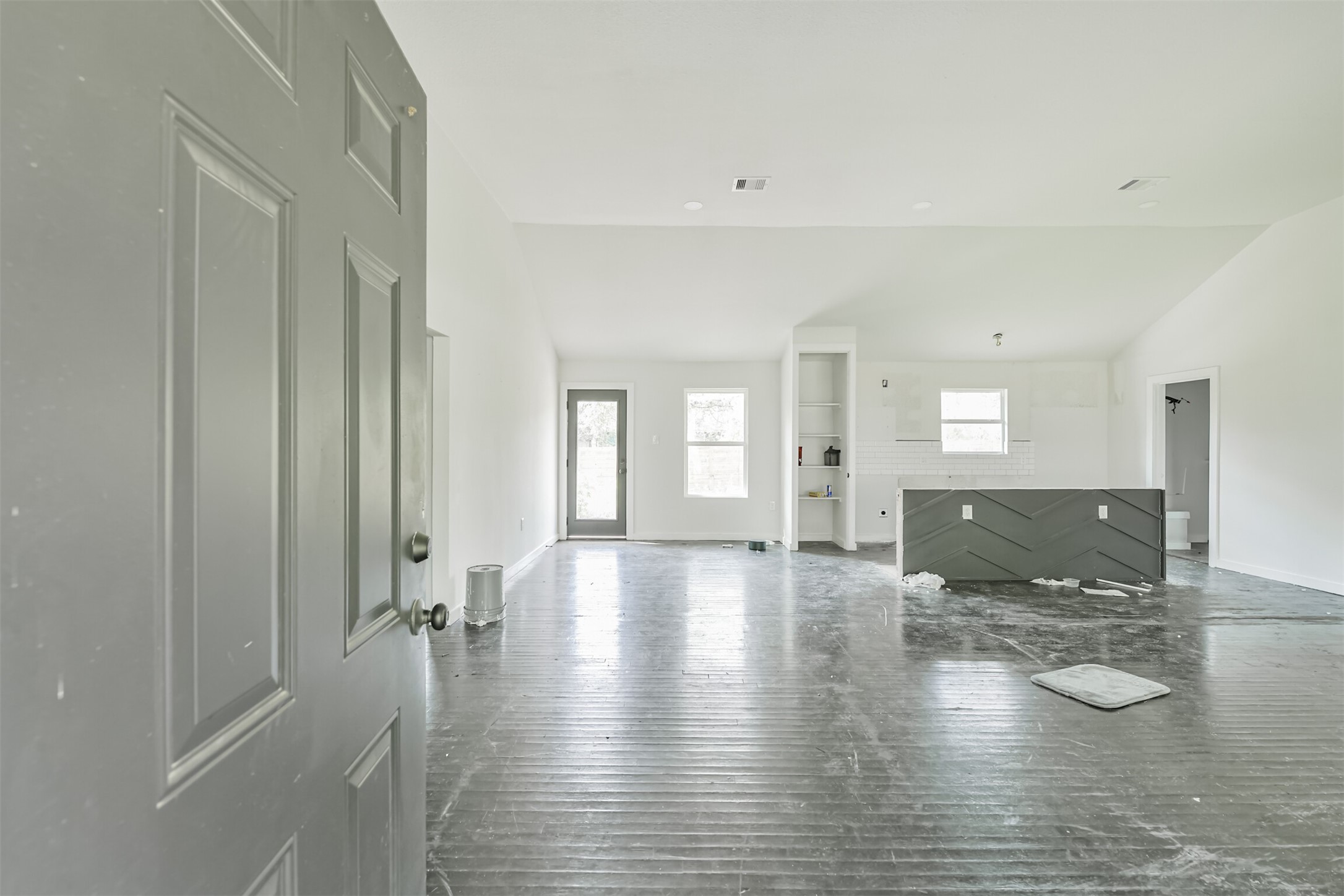 209 Leago Street Houston, TX 77022 - Photo 3 of 24 This photo shows a spacious, unfinished open-concept living area with large windows and a modern kitchen island. The room has high ceilings, white walls, and a door leading outside, offering a bright and airy atmosphere.