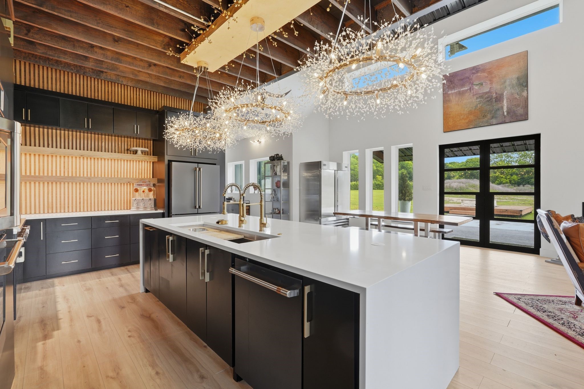 2743 Bobville Road Montgomery, TX 77316 - Photo 20 of 50 This modern kitchen features elegant black cabinetry, a spacious white island with dual sinks, and stunning decorative lighting. High ceilings with exposed beams, large windows, and glass doors provide ample natural light and views of the greenery outside. Perfect for entertaining, it seamlessly connects to a dining area with a sleek, open design.