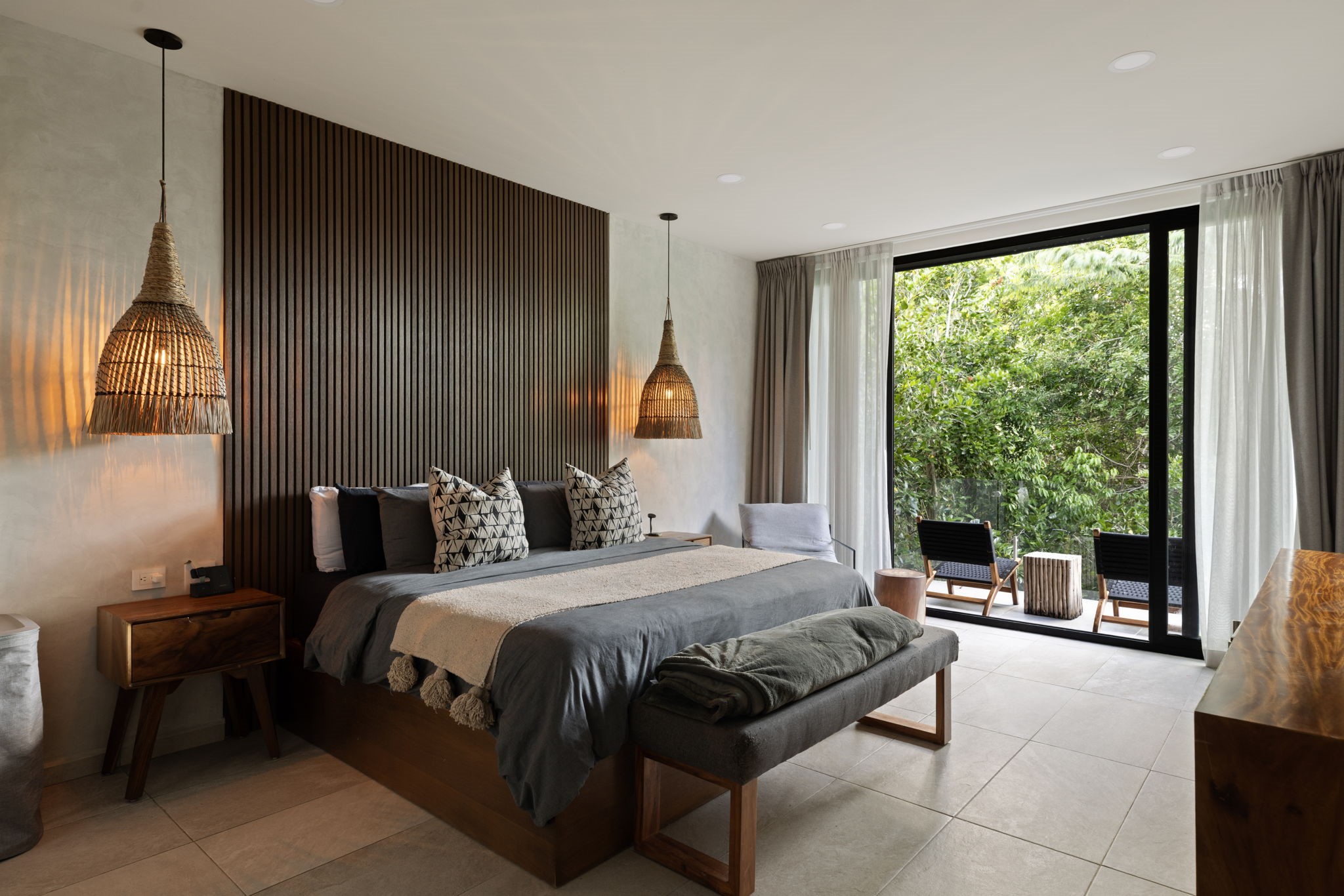 6 Country Club, Unit 4 Nome, TX 77629 - Photo 20 of 45 This spectacular primary suite serves as a luxurious, eco-conscious sanctuary, beautifully marrying refined architectural details with captivating views of the surrounding jungle canopy. The design highlights a rich, floor-to-ceiling wood-slat accent wall and twin woven-rattan basket pendant lights, while oversized glass sliding doors open directly to a secluded balcony oasis.