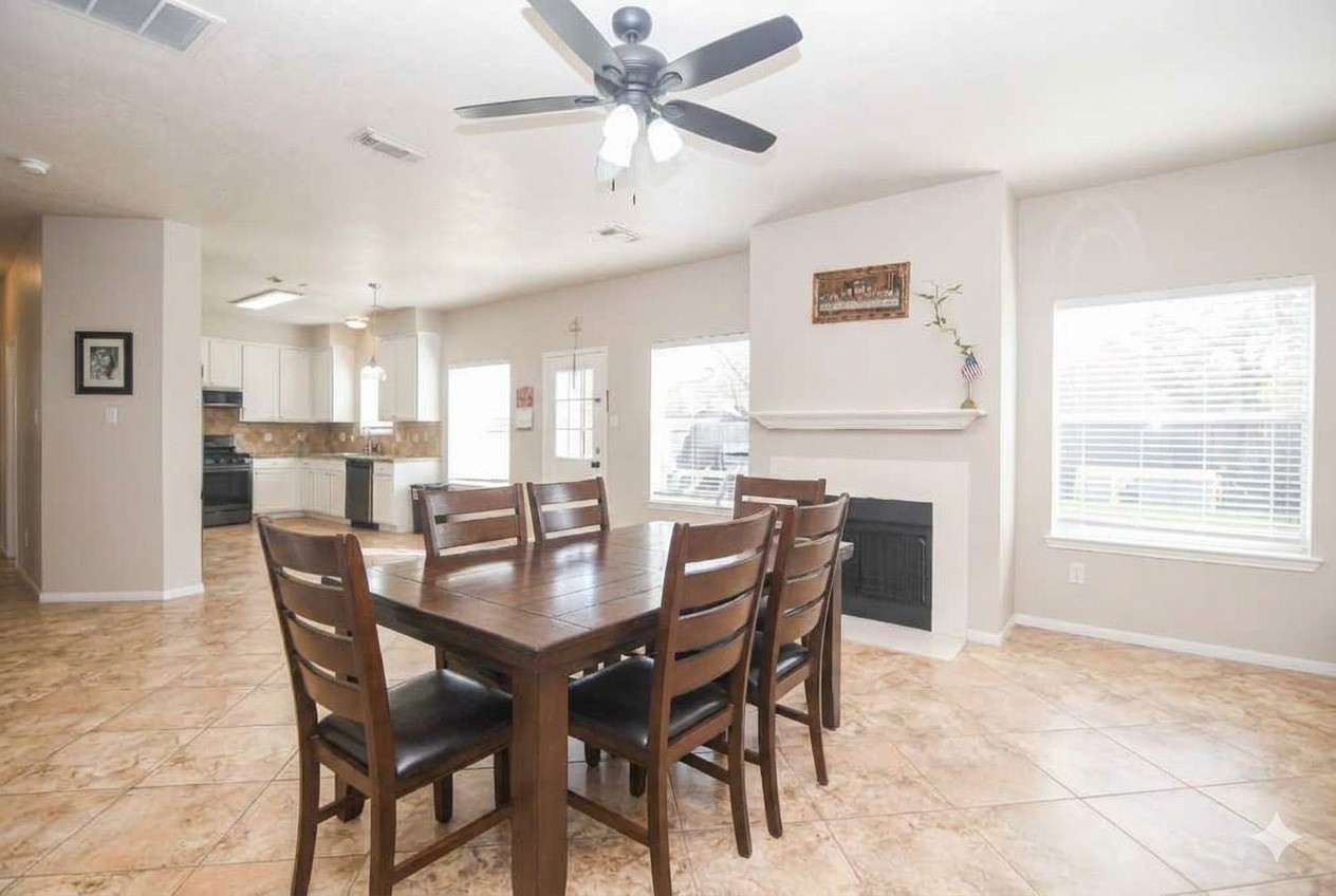 14407 Andrea Way Lane Houston, TX 77083 - Photo 14 of 29 This photo shows a bright, open-concept dining and kitchen area with tiled floors. The dining area features a ceiling fan and a cozy fireplace, while the kitchen offers modern appliances and ample natural light from large windows.