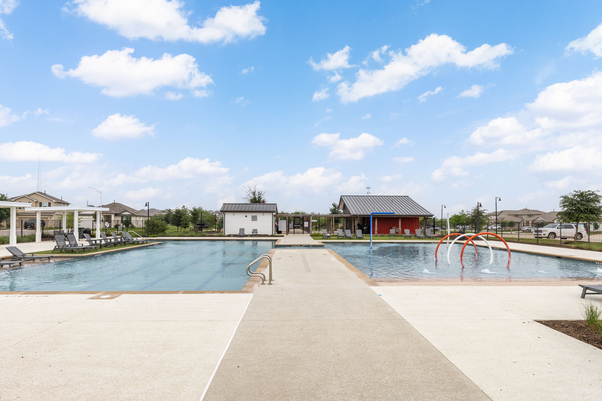 281 Jayden Drive Kyle, TX 78640 - Photo 33 of 36 Community pool area featuring a large swimming pool, a separate splash pad with water features, a shaded lounging area, a red-sided pavilion, and a white auxiliary building