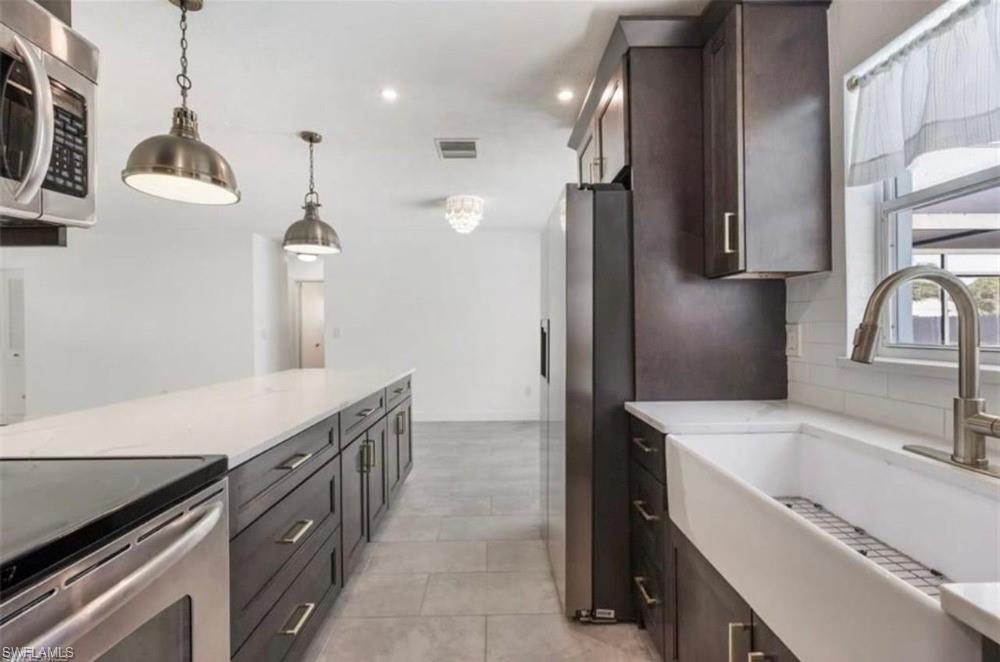 1948 46th Street Southwest Naples, FL 34116 - Photo 5 of 14 Modern kitchen featuring dark wood cabinetry, stainless steel appliances, a large center island with a light-toned countertop, a farmhouse sink, and contemporary pendant lighting