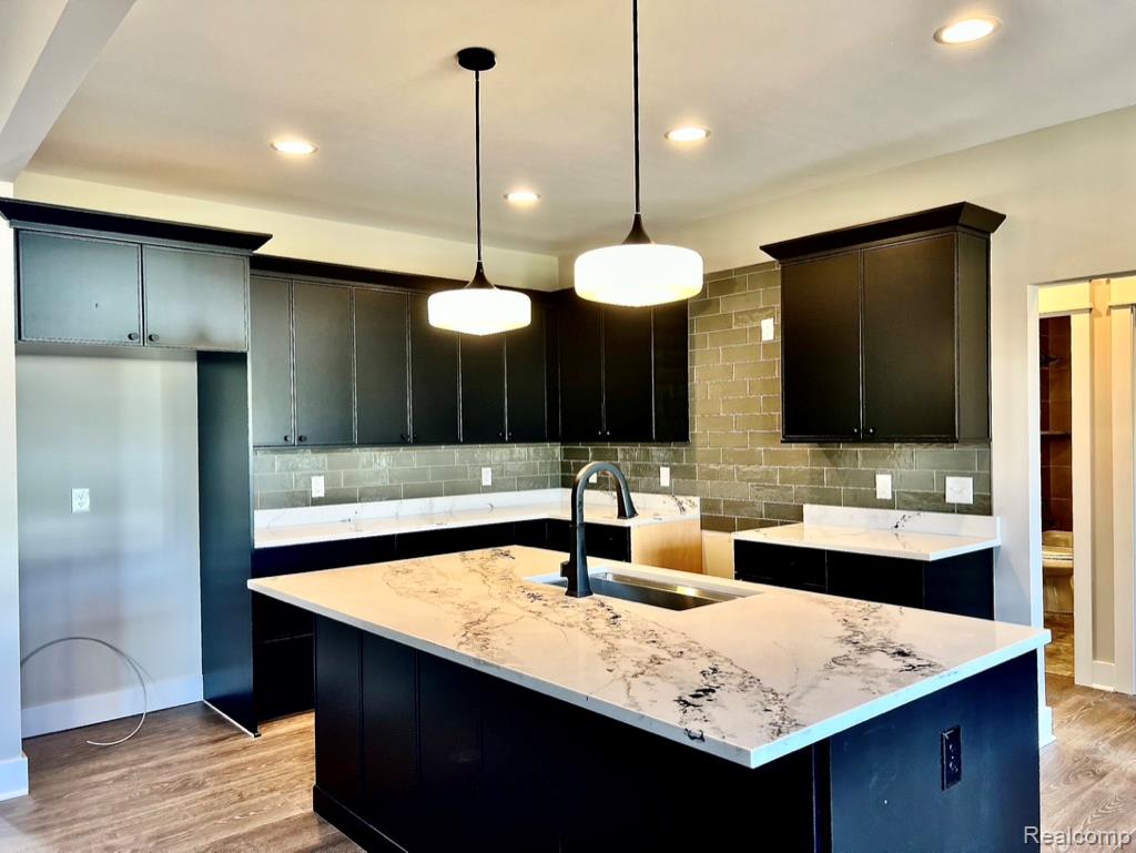 5382 Spring Meadows Way Linden, MI 48451 - Photo 9 of 20 Kitchen featuring a kitchen island with sink, light stone counters, light wood-style flooring, pendant lighting, and decorative backsplash