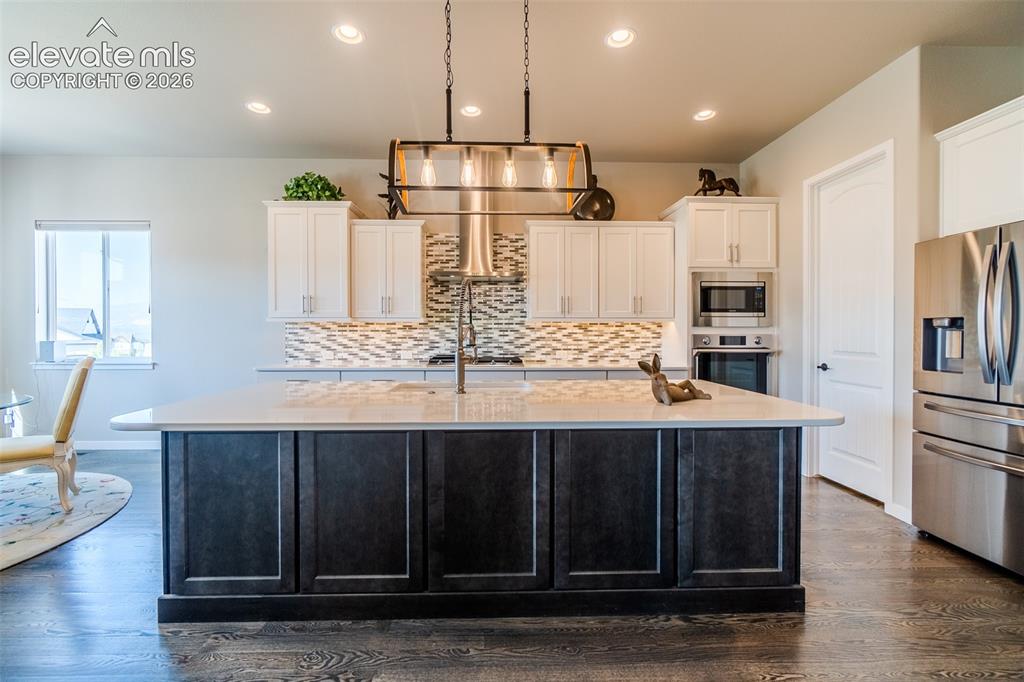 1061 Native Dancer Terrace Colorado Springs, CO 80921 - Photo 11 of 50 Kitchen featuring an expansive center island with a light-toned countertop, a stylish linear pendant light fixture, stainless steel appliances, a mosaic tile backsplash, and wood-finish flooring