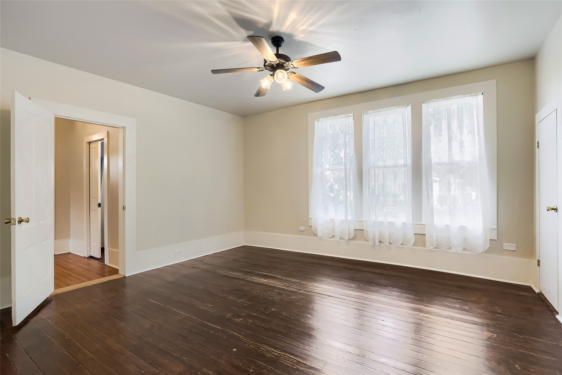 208 North Ave B Elgin, TX 78621 - Photo 20 of 29 This spacious room features polished dark wood floors, large windows with sheer curtains for natural light, and a ceiling fan for comfort. The neutral walls and white trim create a clean, bright atmosphere, while the doorways lead to additional living spaces.