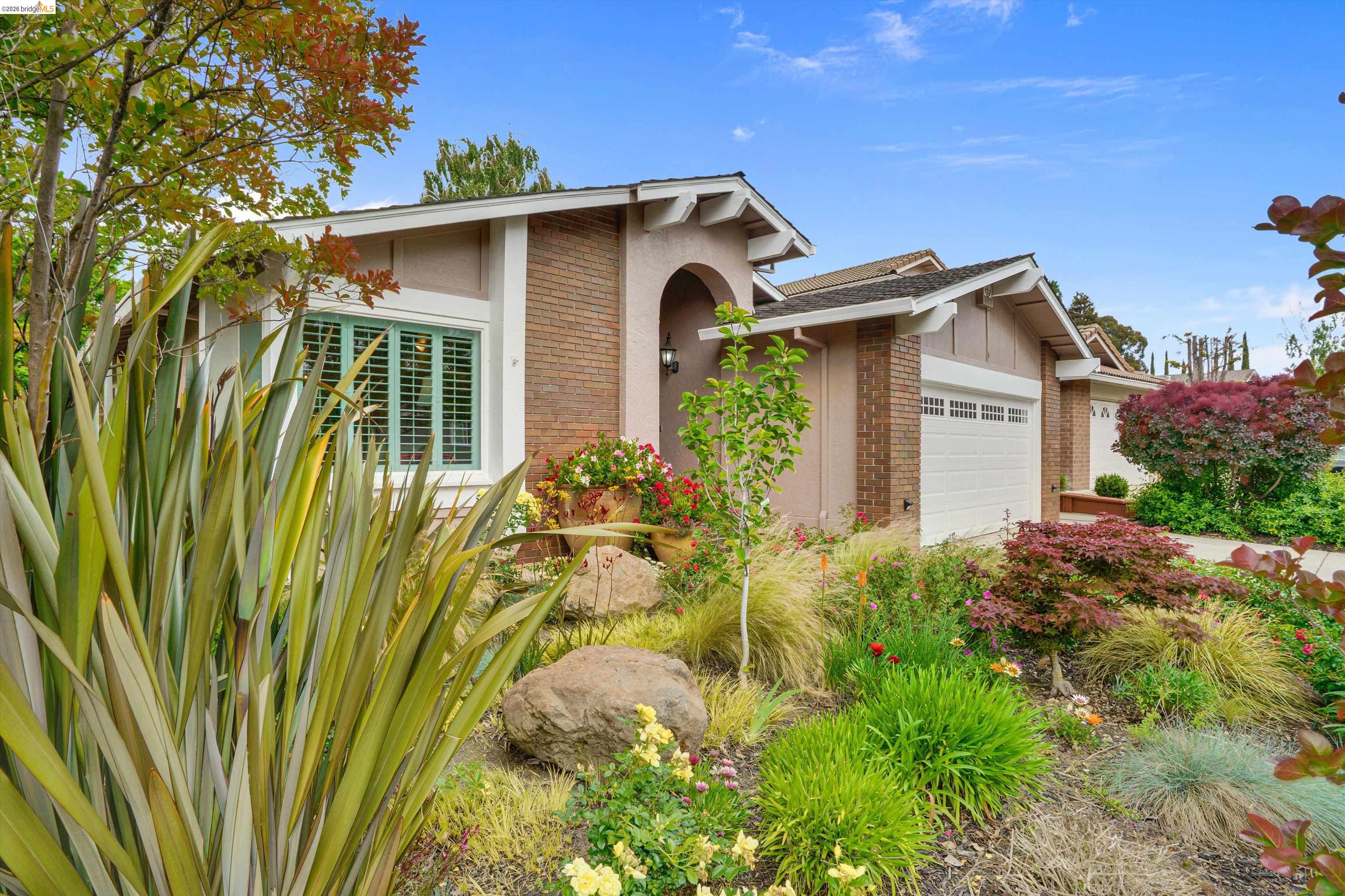 Lush, colorful landscaping frames this property, featuring a brick facade with stucco accents, an arched entryway, a large window with green shutters, and a two-car garage with white doors
