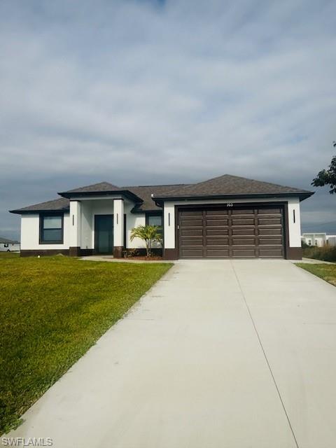 165 Patio Street Lehigh Acres, FL 33974 - Photo 2 of 34 Prairie-style home featuring a front lawn, concrete driveway, a garage, roof with shingles, and stucco siding