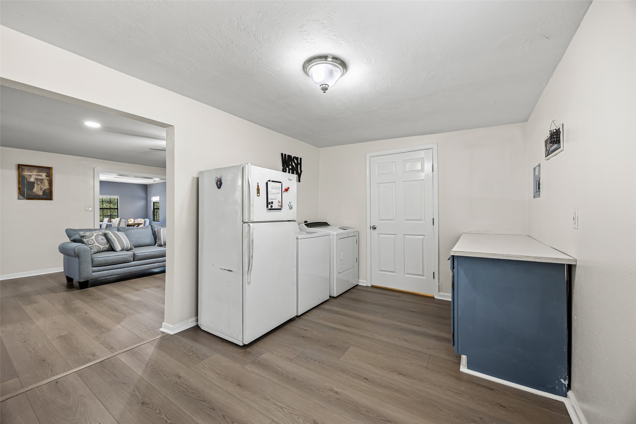15080 Usher Lane Splendora, TX 77372 - Photo 39 of 44 This photo showcases a bright, spacious laundry room. It features light wood flooring and a ceiling light. An open doorway leads to a cozy living area with a blue couch and natural light from large windows.
