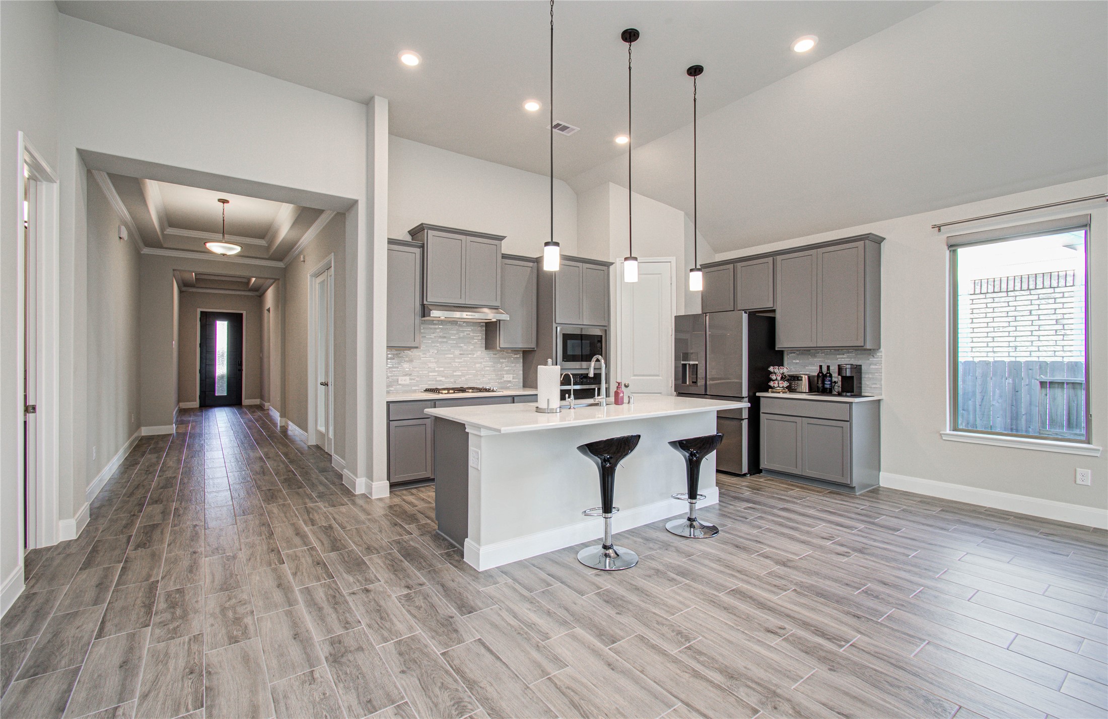17344 Grand Canyon Road Conroe, TX 77302 - Photo 15 of 49 This modern kitchen features sleek gray cabinetry, a large island with seating, quartz contertops, and pendant lighting. It opens into a spacious hallway with wood-look tile flooring and high ceilings, creating an airy and inviting atmosphere.
