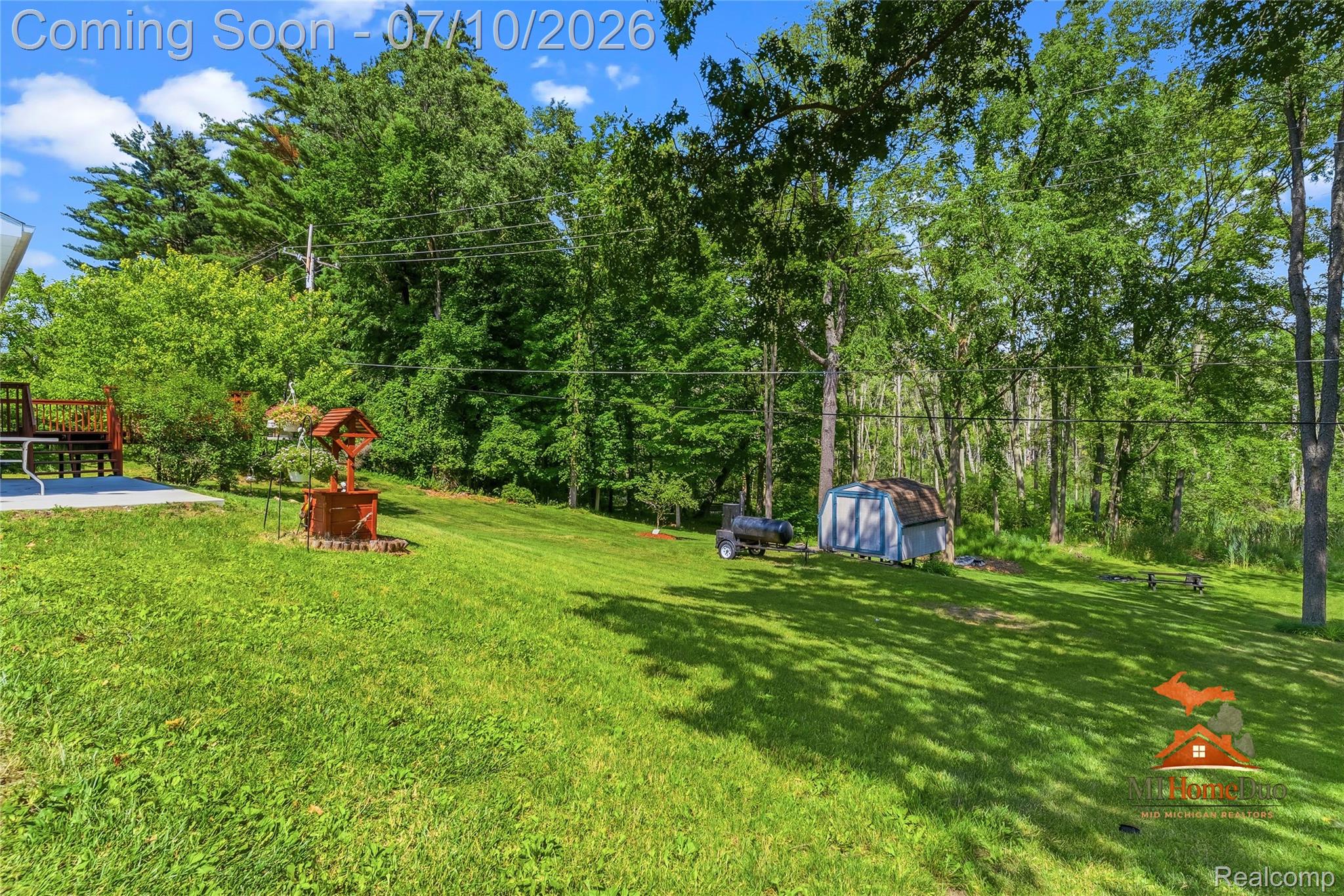 11231 Mcgregor Road Pinckney, MI 48169 - Photo 27 of 37 Expansive green lawn framed by mature trees, featuring a concrete patio, wood deck with railing, and a storage shed