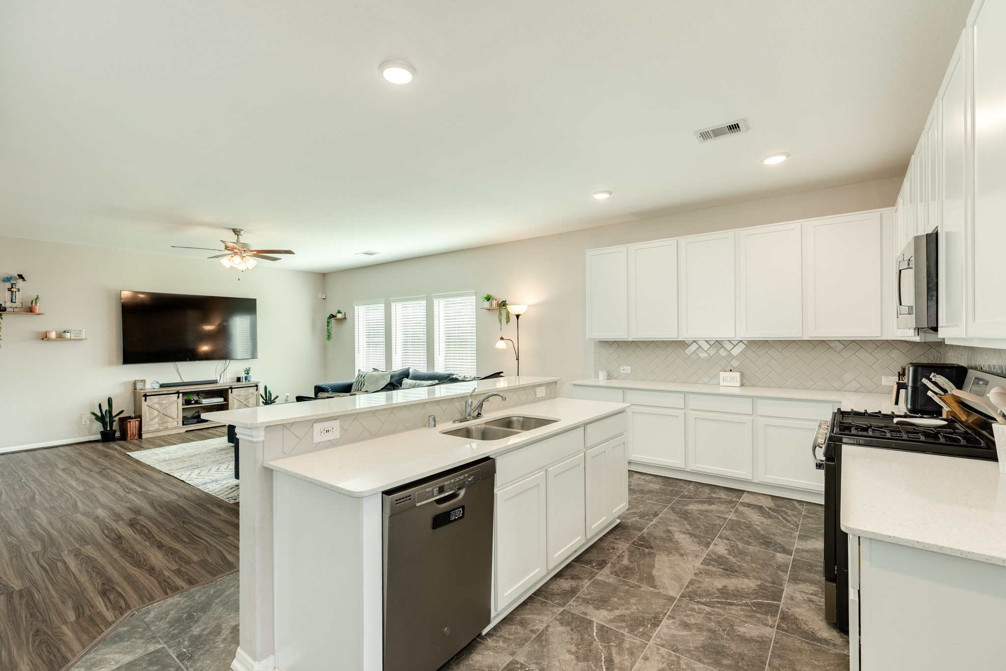 2446 Palisade Crst Drive Rosharon, TX 77583 - Photo 10 of 30 This photo shows a modern, open-concept living space with a spacious kitchen featuring white cabinets, a large island with a sink, and stainless steel appliances. The living area has wood look flooring, a mounted TV, and ample natural light from multiple windows.