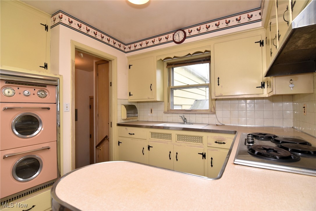 9421 David Road Cleveland, OH 44125 - Photo 15 of 36 Kitchen featuring under cabinet range hood, oven, stainless steel stovetop, decorative backsplash, and estacked washer and dryer