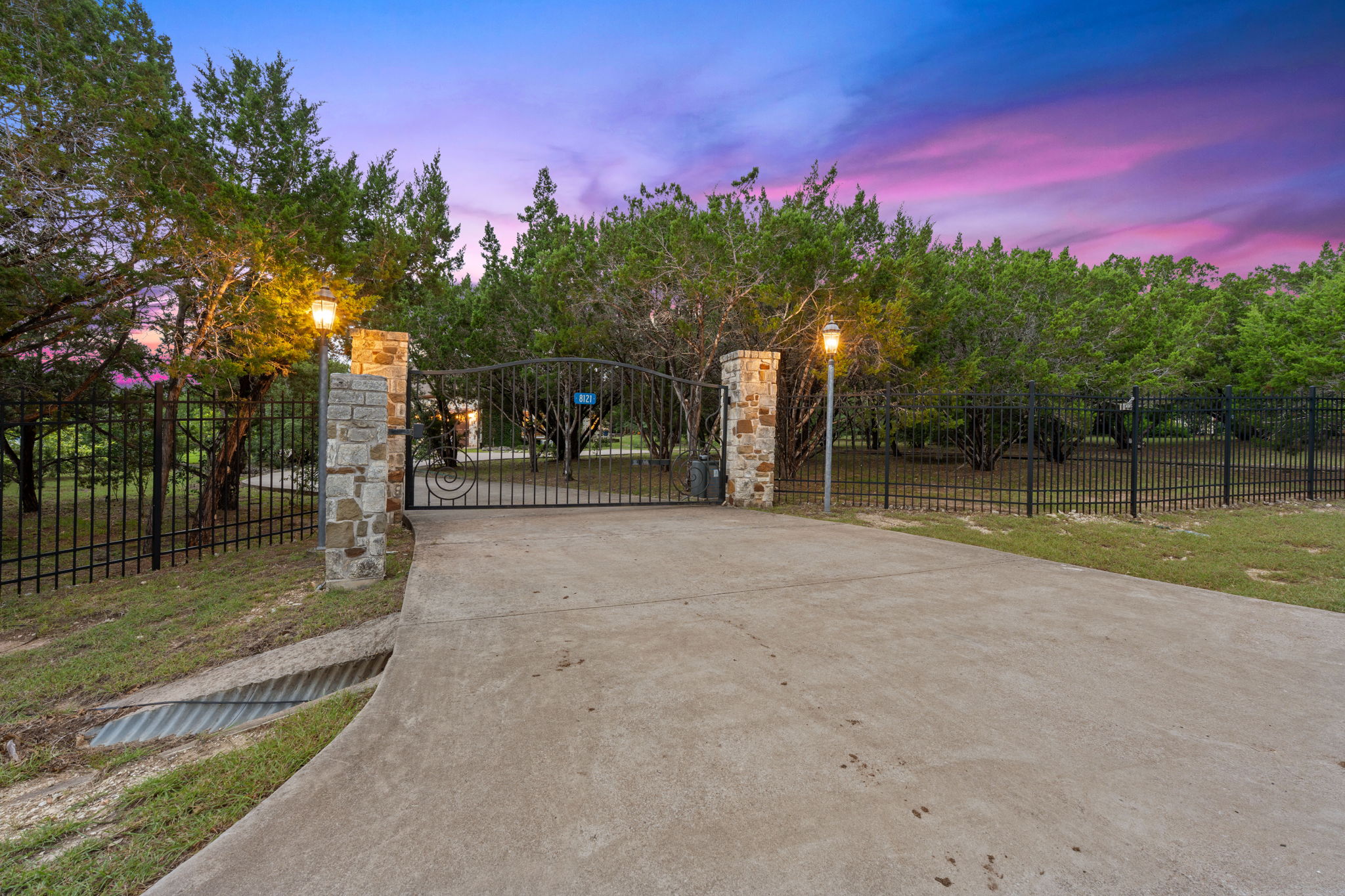 8121 Bluff Trail Austin, TX 78737 - Photo 2 of 40 Secure entry through a wrought iron gate with stone pillars and illuminating post lamps, framed by natural trees.