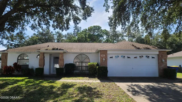a view of a white house in front of a yard with plants and large trees