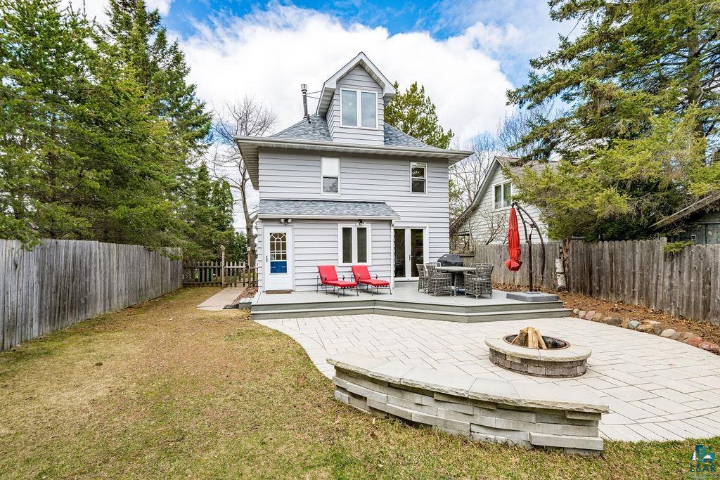 Rear exterior featuring a new deck, a large paver patio with a built-in stone fire pit, extensive lawn space, and mature perimeter trees leading to the beach.