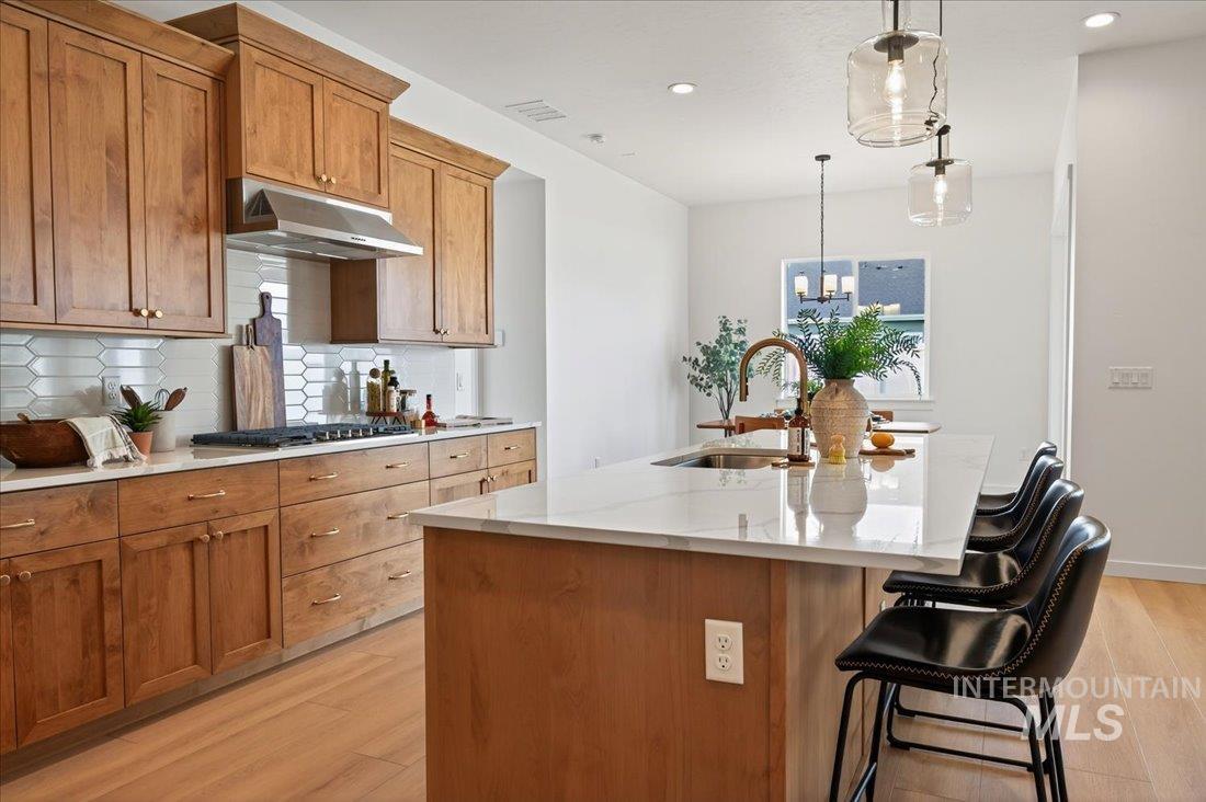 14675 Serenity Pt Street Caldwell, ID 83607 - Photo 7 of 14 Kitchen with brown cabinetry, a kitchen island with sink, light stone countertops, under cabinet range hood, and light wood-type flooring