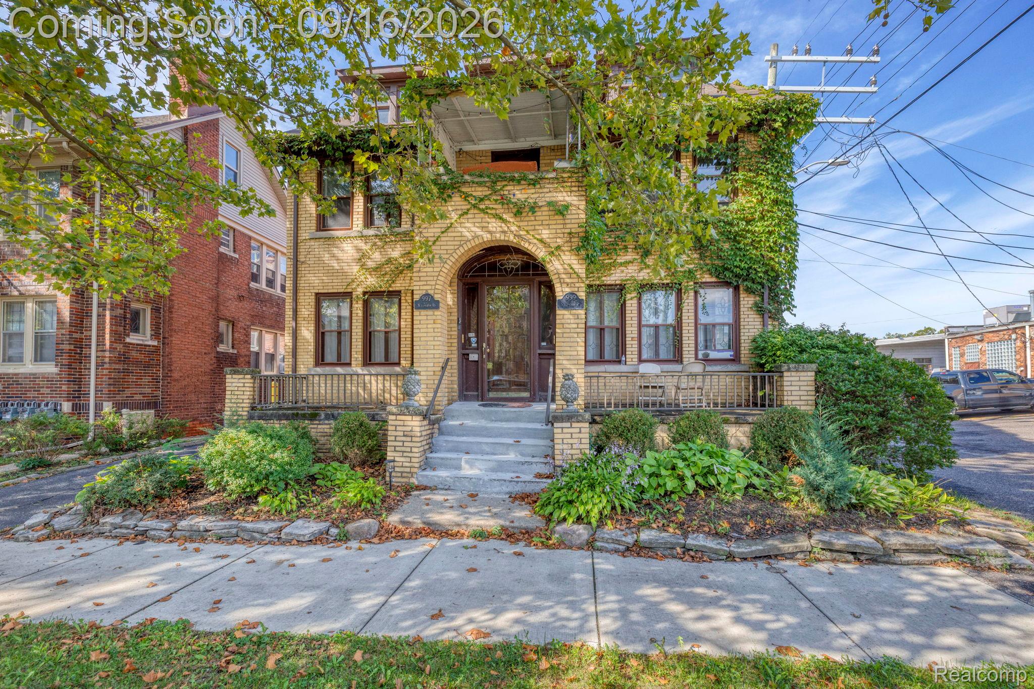 Brick exterior with a covered front porch, arched entryway, mature landscaping, and an upper-level balcony