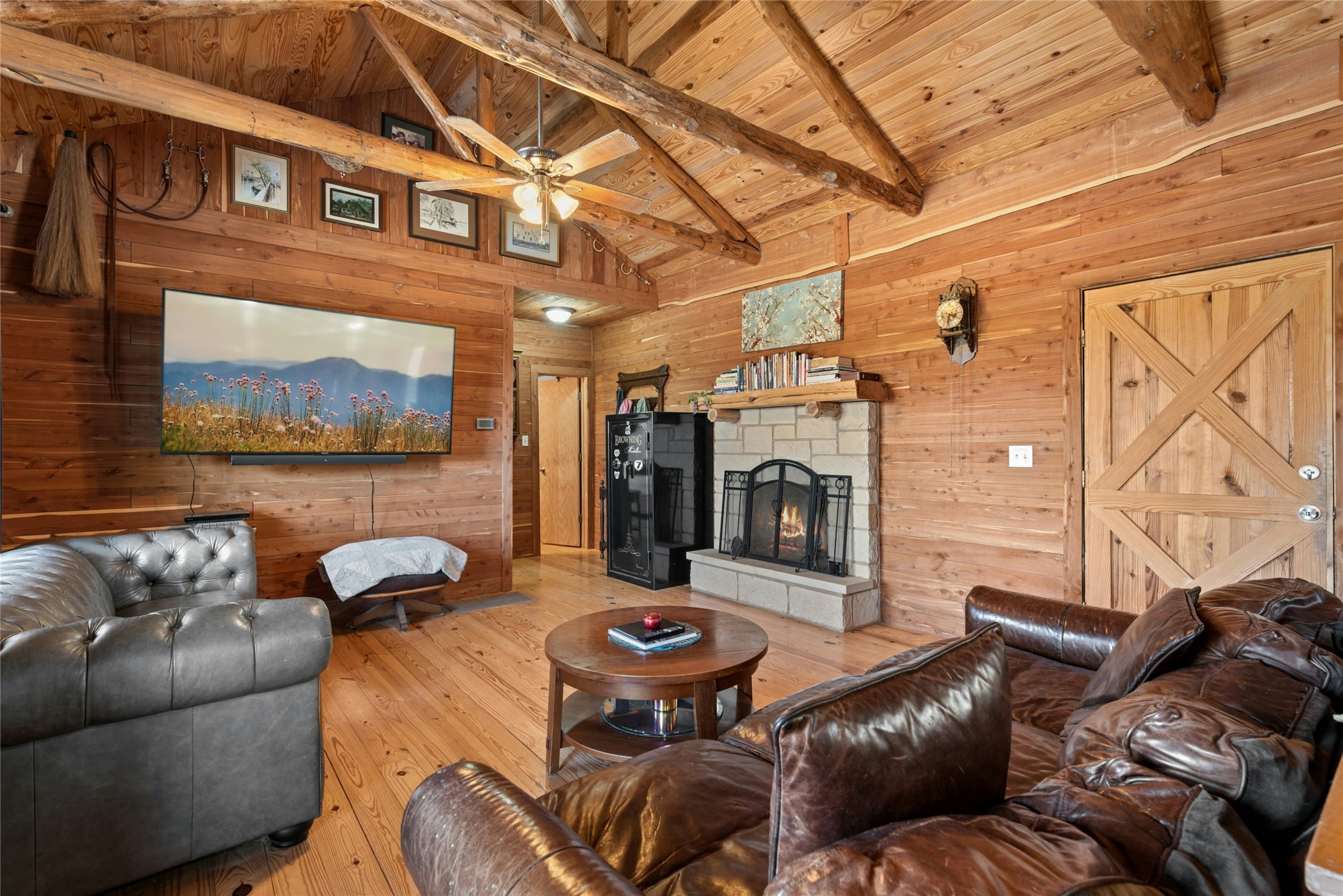 496 Middleton Road Spicewood, TX 78669 - Photo 10 of 40 Living area featuring natural wood paneling, exposed beam vaulted ceiling, a stone-surround fireplace with hearth, wood-finish flooring, and a ceiling fan