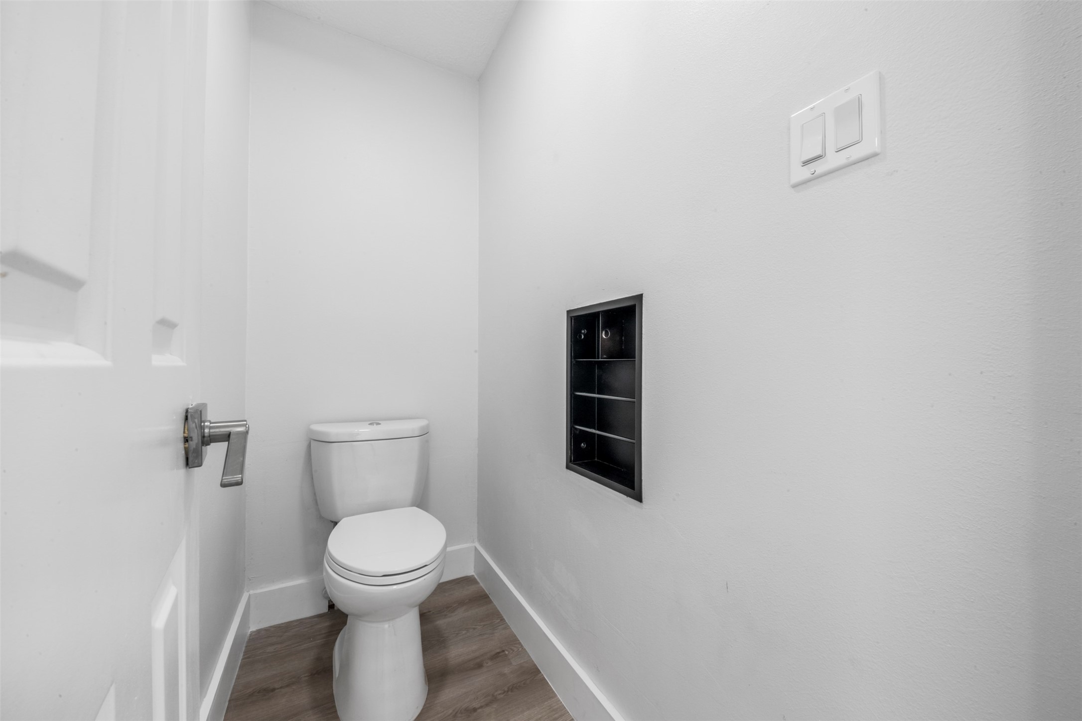 9543 Enstone Circle Spring, TX 77379 - Photo 33 of 50 This photo shows a small, modern bathroom featuring a toilet, light gray walls, and wood-style flooring. A recessed shelf is built into the wall, adding functional storage space.