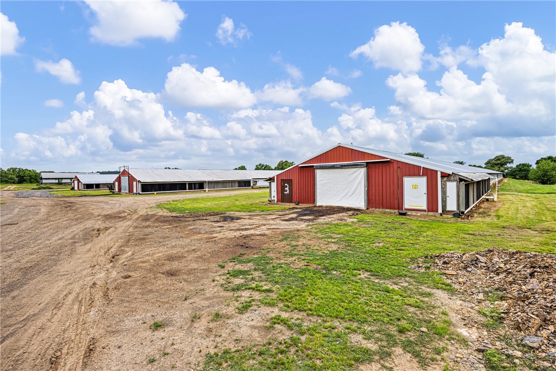 Multiple red-sided agricultural buildings featuring white metal roofing and large roll-up doors, set against a backdrop of green fields and a blue sky with clouds