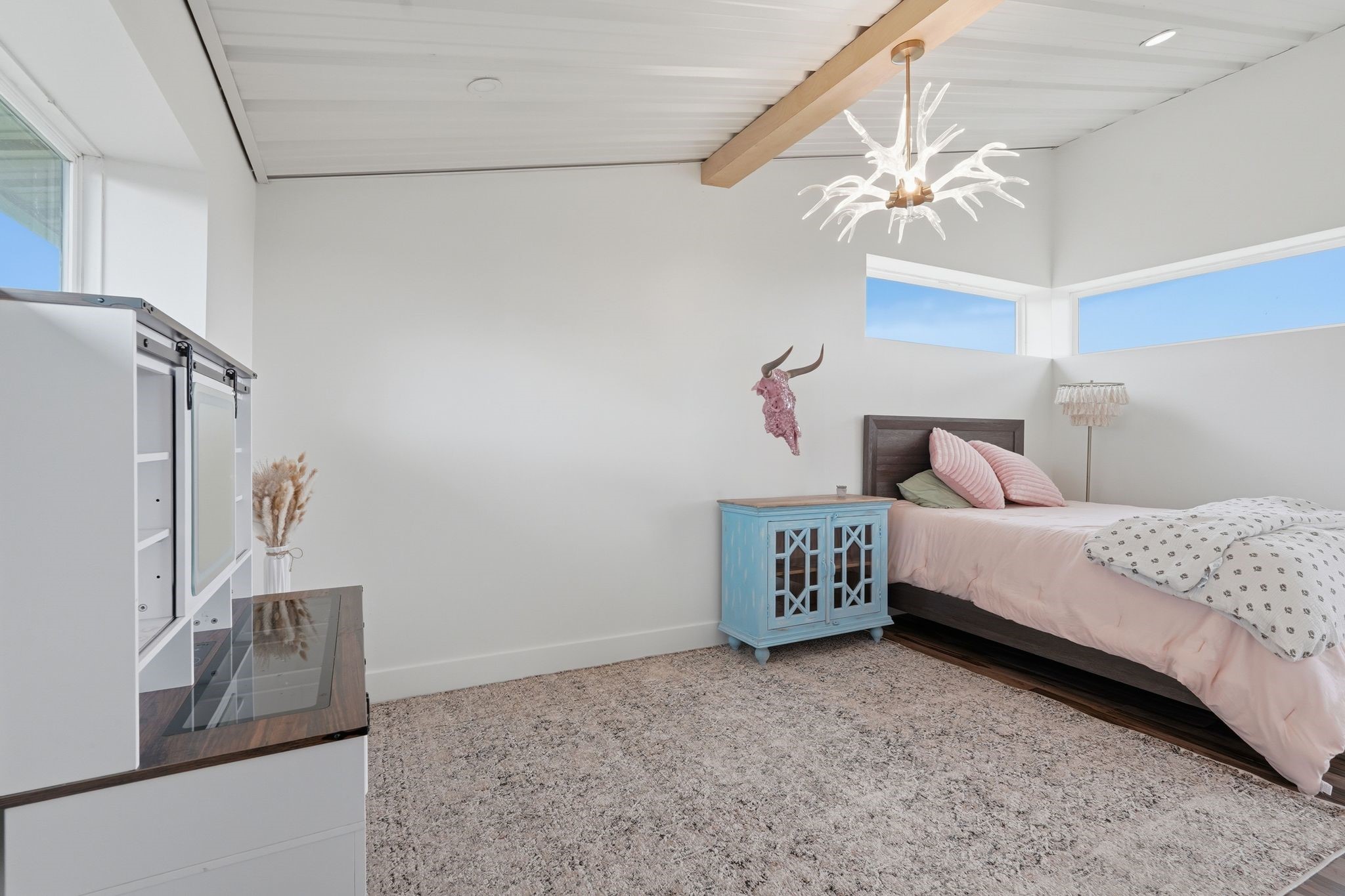2743 Bobville Road Montgomery, TX 77316 - Photo 38 of 50 This bedroom features a modern, minimalist design with white walls and a carpeted floor. It includes a single bed with pink bedding, a unique antler-style ceiling light, and a blue bedside cabinet. Natural light streams through high, horizontal windows, creating a bright and airy atmosphere.