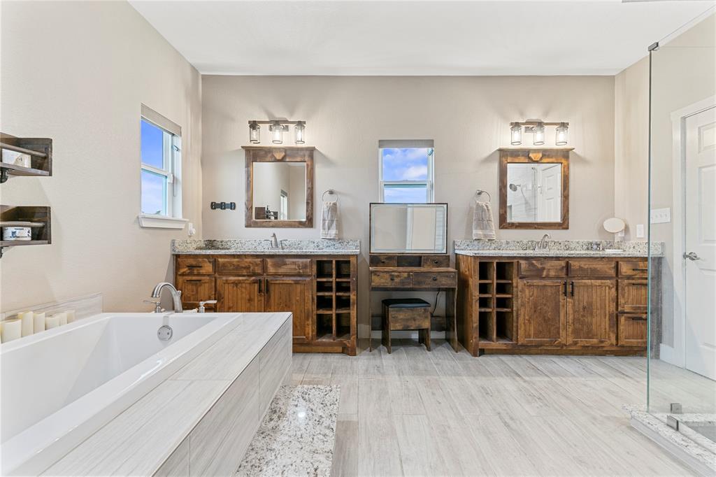 139 Kriston Lane Springtown, TX 76082 - Photo 24 of 39 Spacious bath featuring dual wood-finish vanities with granite countertops, a built-in makeup vanity, a freestanding soaking tub with tile surround, and a glass-enclosed shower