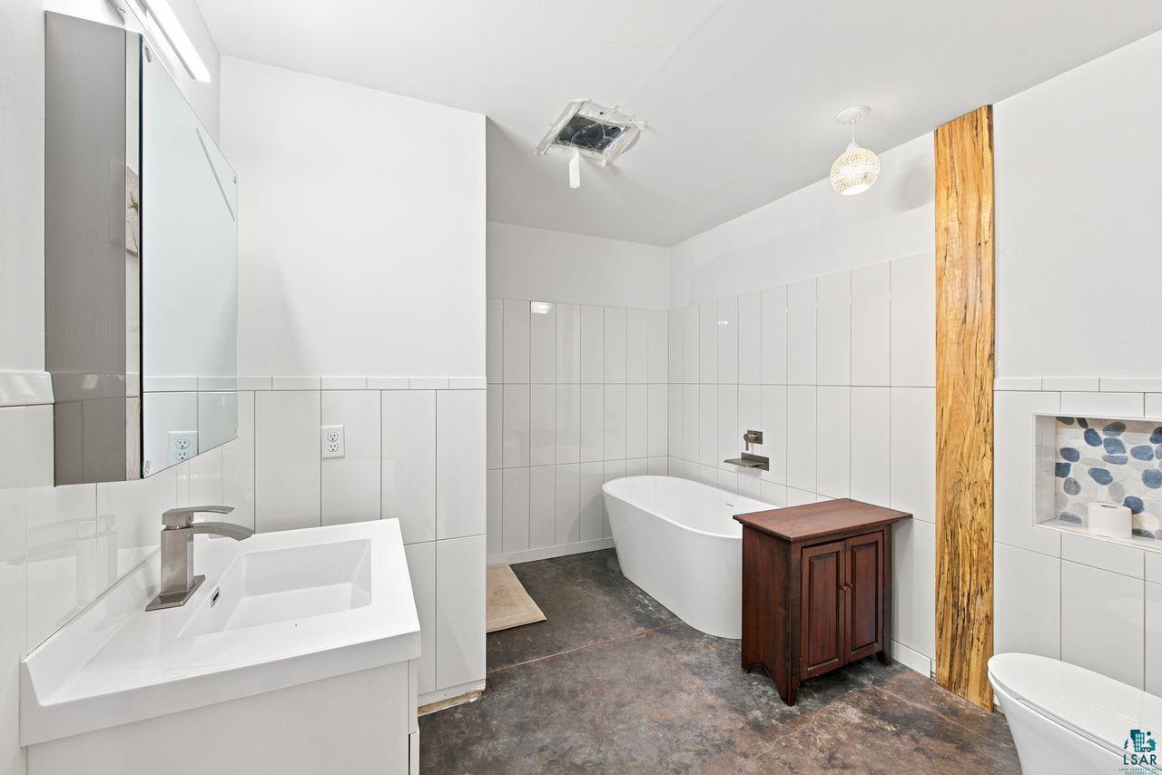 5400 Eightmile Lake Road Iron River, WI 54847 - Photo 15 of 37 Contemporary bathroom featuring a freestanding tub, white wall tiling, a vanity with an integrated sink, a mirrored medicine cabinet, and a rustic wood accent column