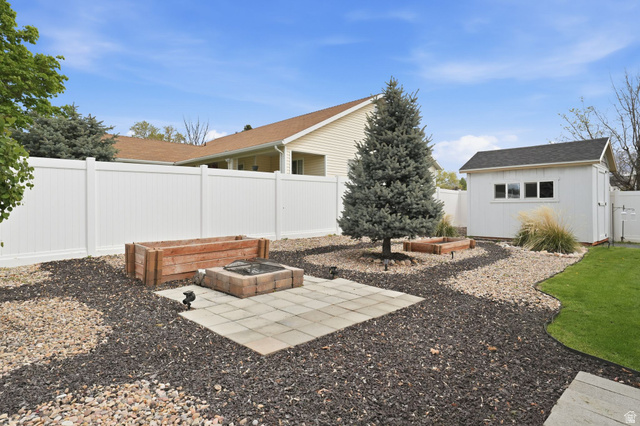 2190 West 1450th Street North Layton, UT 84041 - Photo 6 of 11 Expansive backyard featuring a white vinyl privacy fence, a detached storage shed with multiple windows, a mature evergreen tree, and a paved patio area with a built-in fire pit