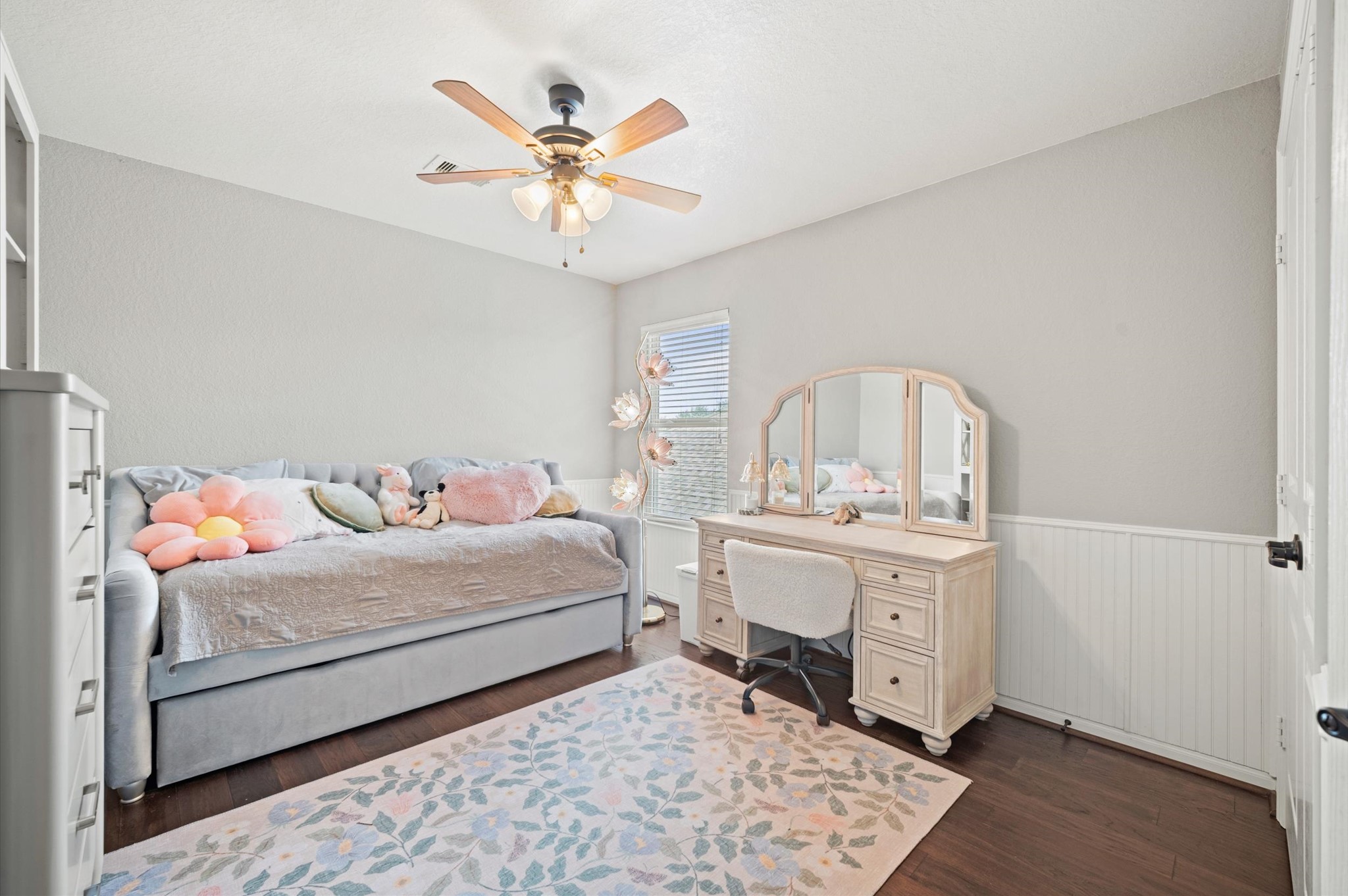 9507 Empress Crossing Drive Spring, TX 77379 - Photo 36 of 50 This cozy bedroom features a soft color palette with a daybed adorned with plush pillows, a ceiling fan, and a charming vanity set. Natural light flows in through a window, highlighting the floral rug and elegant hardwood flooring. Perfect for a relaxing retreat.
